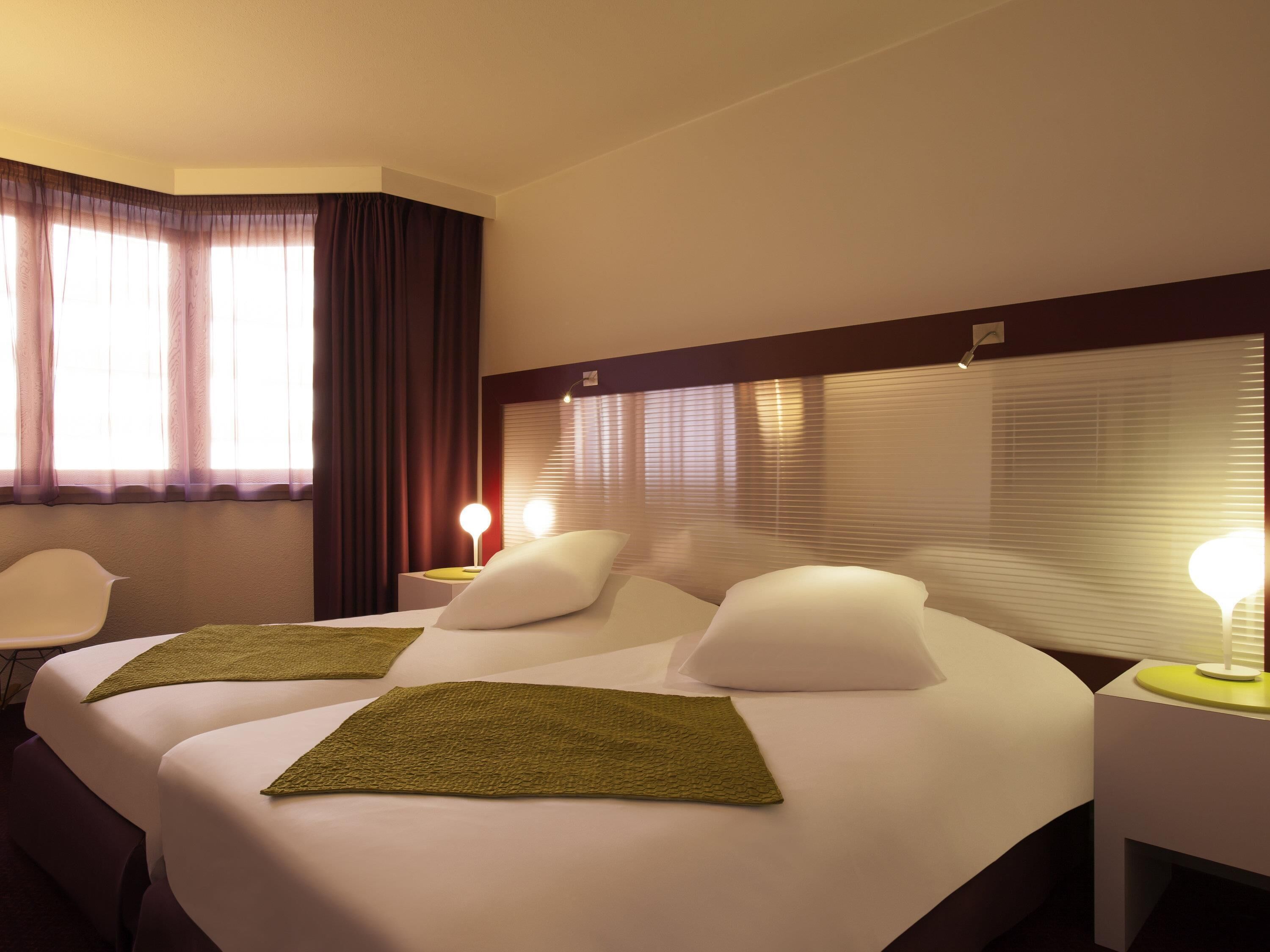 Une chambre d’hôtel avec un grand lit, deux tables de chevet, une chaise et une fenêtre avec des rideaux.