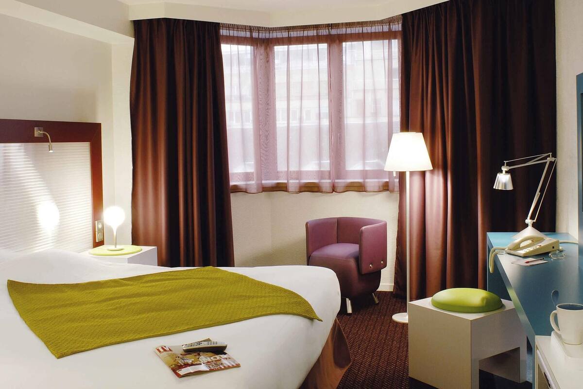 Une chambre d’hôtel avec un lit, un bureau doté d’une lampe, une chaise et une fenêtre avec des rideaux.