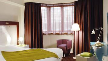 Una habitación de hotel con cama, escritorio con lámpara, silla y ventana con cortinas.