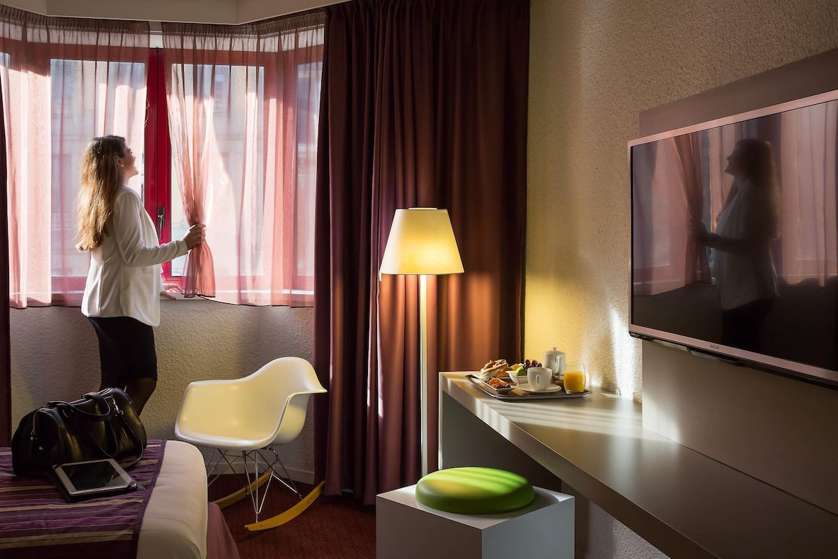 Une chambre d’hôtel avec un lit, une chaise, un bureau avec une lampe, une télévision et une fenêtre avec des rideaux.