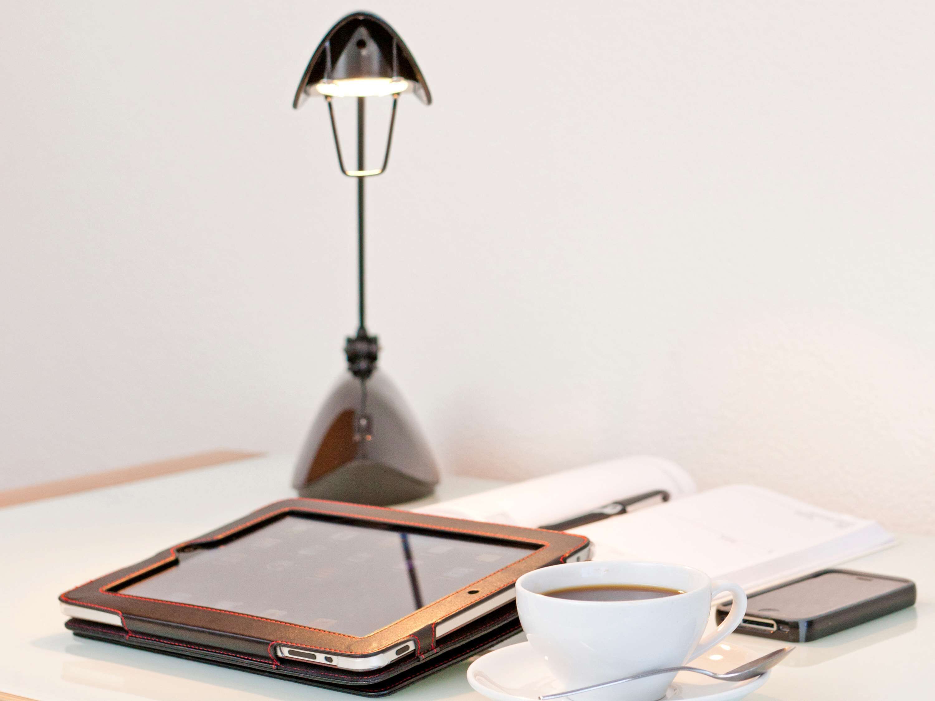 Une tablette avec une housse en cuir, une tasse de café, un smartphone et une lampe de bureau sur une table.