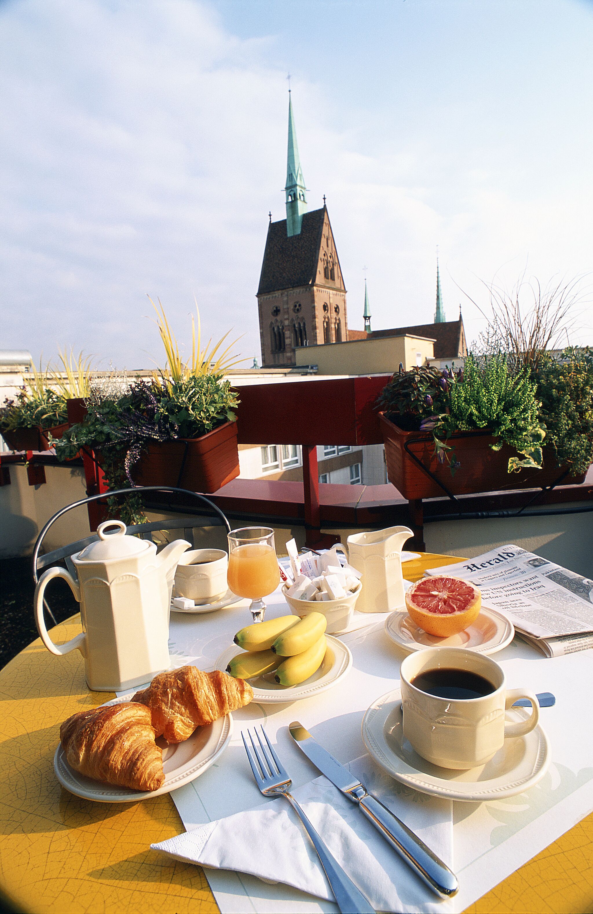 A breakfast setup with croissants, fruit, coffee, and a newspaper on a table with a view of a church spire in the background.