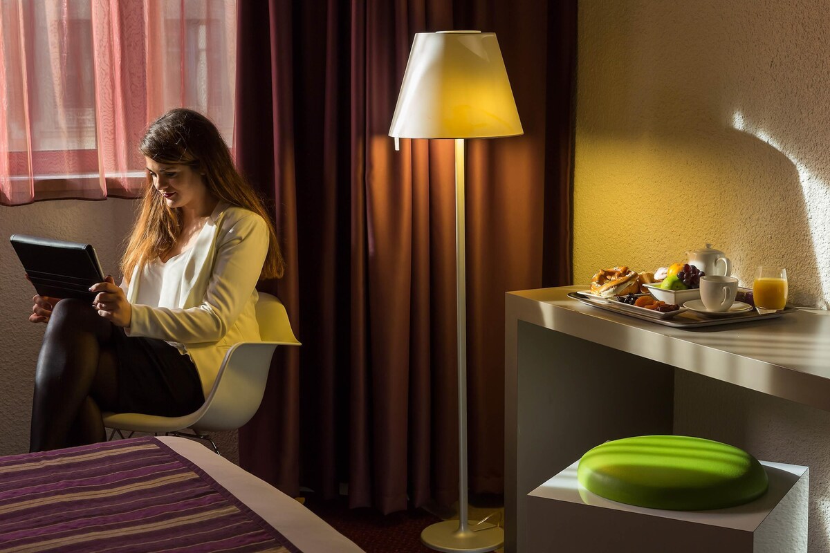 Een hotelkamer met een bed, een bureau met ontbijtartikelen, een lamp, en een persoon die een tablet gebruikt.