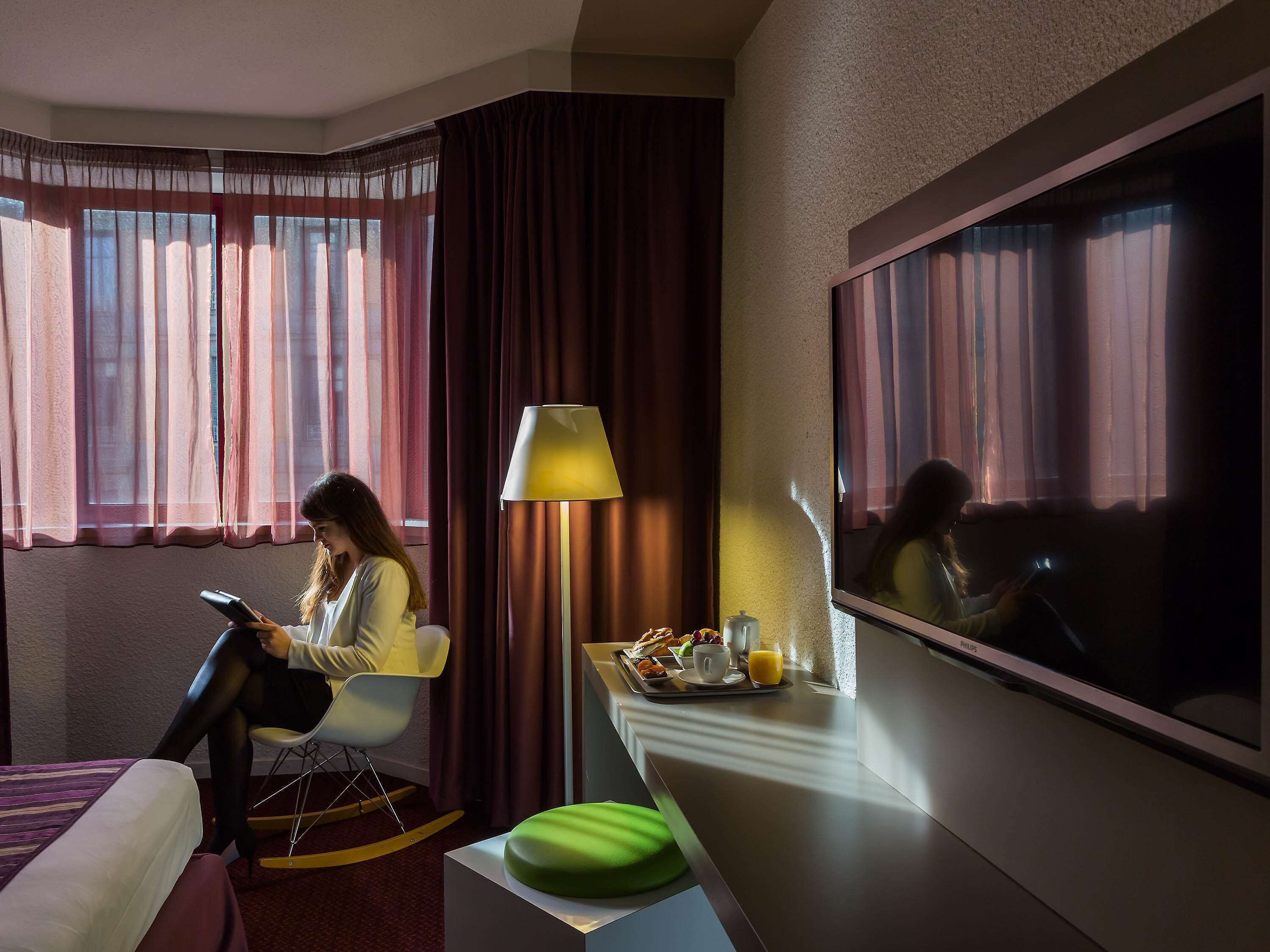 A hotel room with a bed, a chair, a desk with a lamp, a tray with breakfast items, and a large window with curtains.
