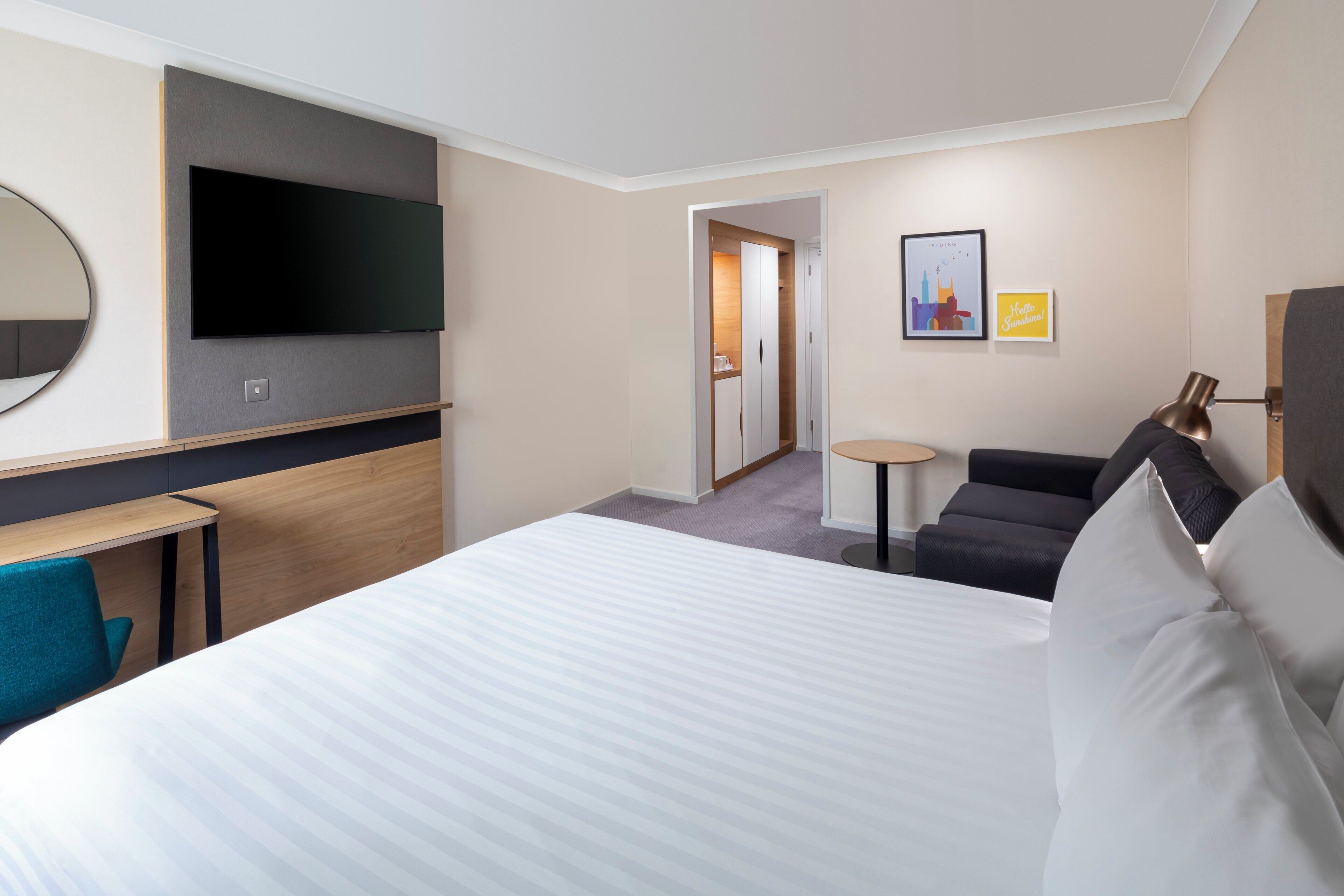 Ein Hotelzimmer mit einem großen Bett, einem an der Wand befestigten Flachbildfernseher, einem kleinen Tisch, einem Sofa und einer Tür zu einem weiteren Raum.