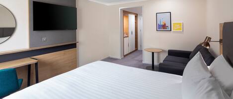 Habitación de hotel con una cama grande, un televisor de pantalla plana montado en la pared, una mesita, un sofá y una puerta que da a otra habitación.