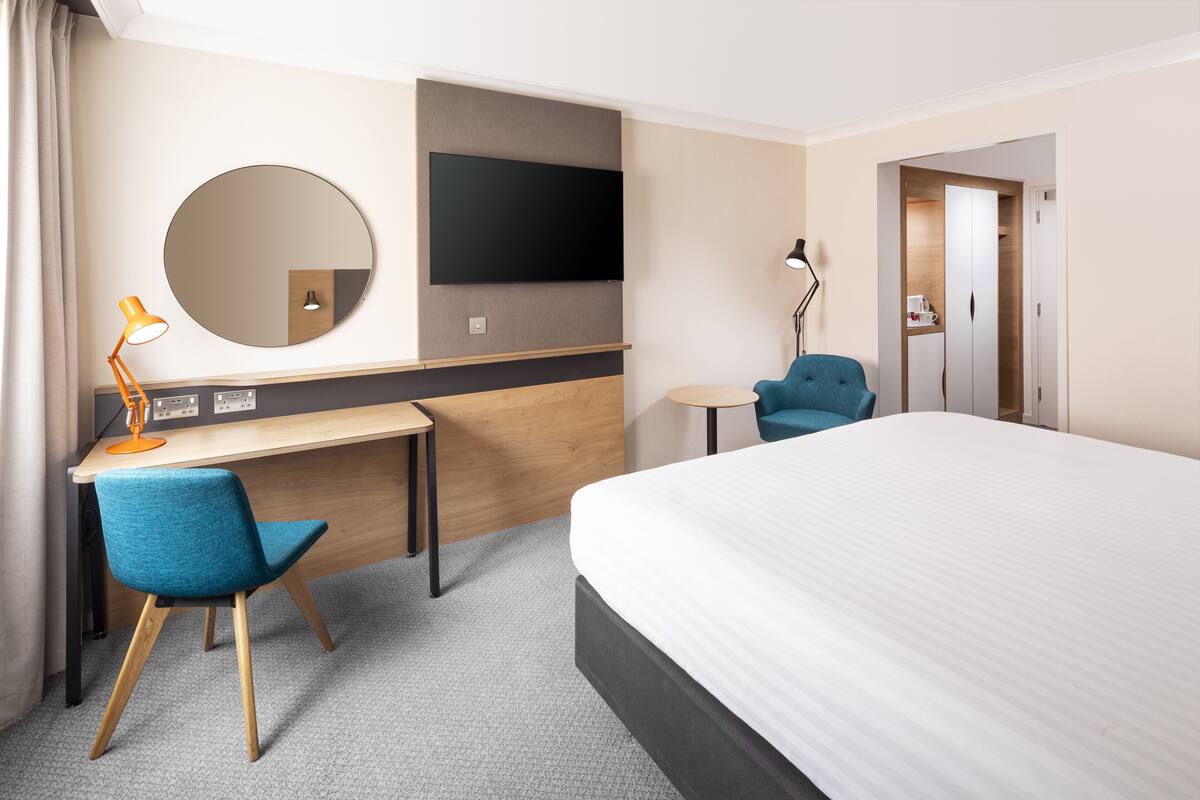 A hotel room with a bed, a desk with a chair, a television, a mirror, and a wardrobe.