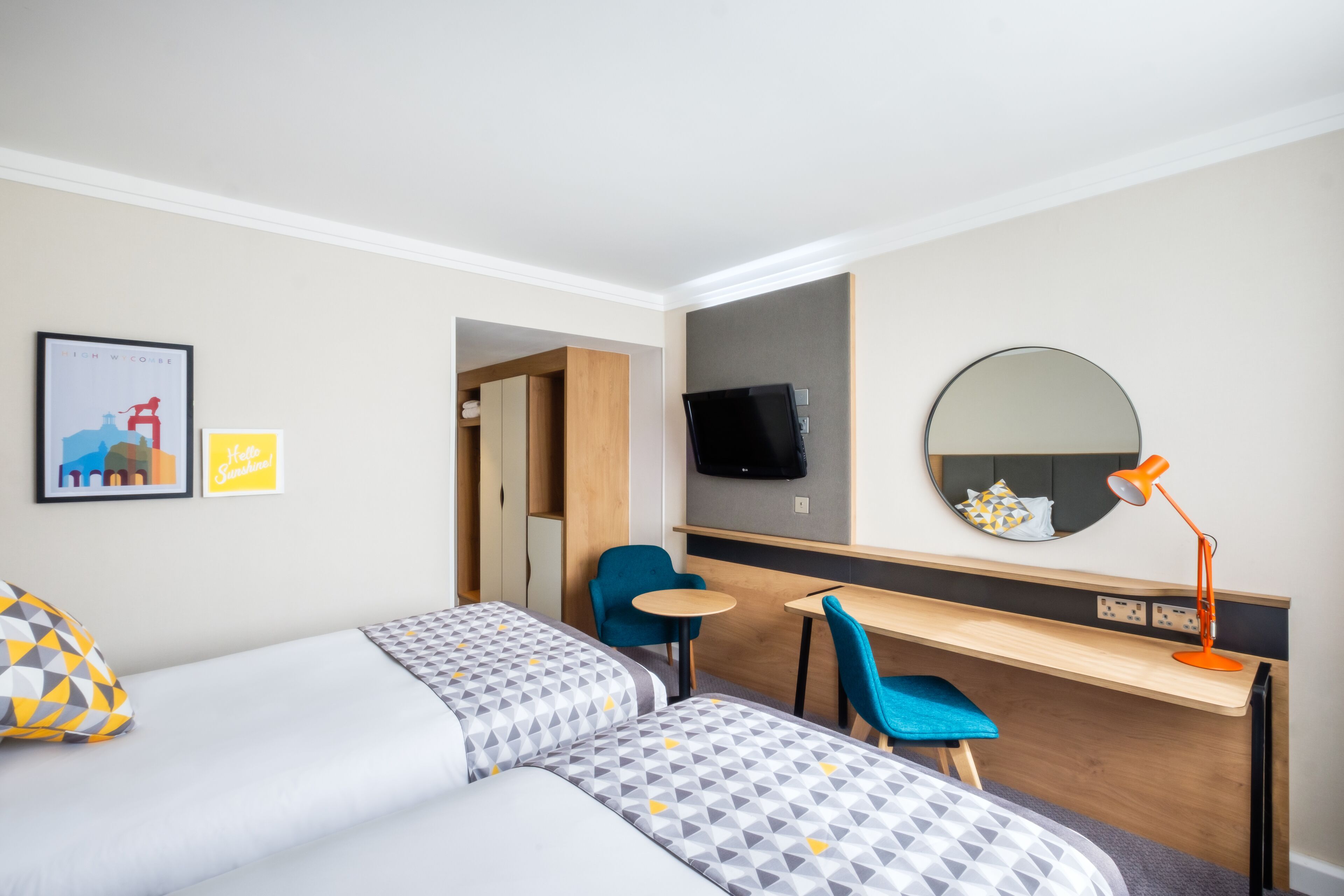 Quarto de hotel com duas camas, uma escrivaninha, uma cadeira, televisão, espelho e uma mesinha.