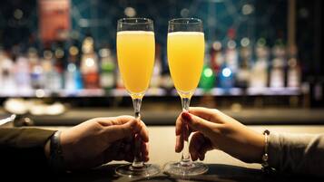 Two hands holding champagne glasses filled with a yellow liquid, likely champagne, at a bar with a blurred background of bottles.
