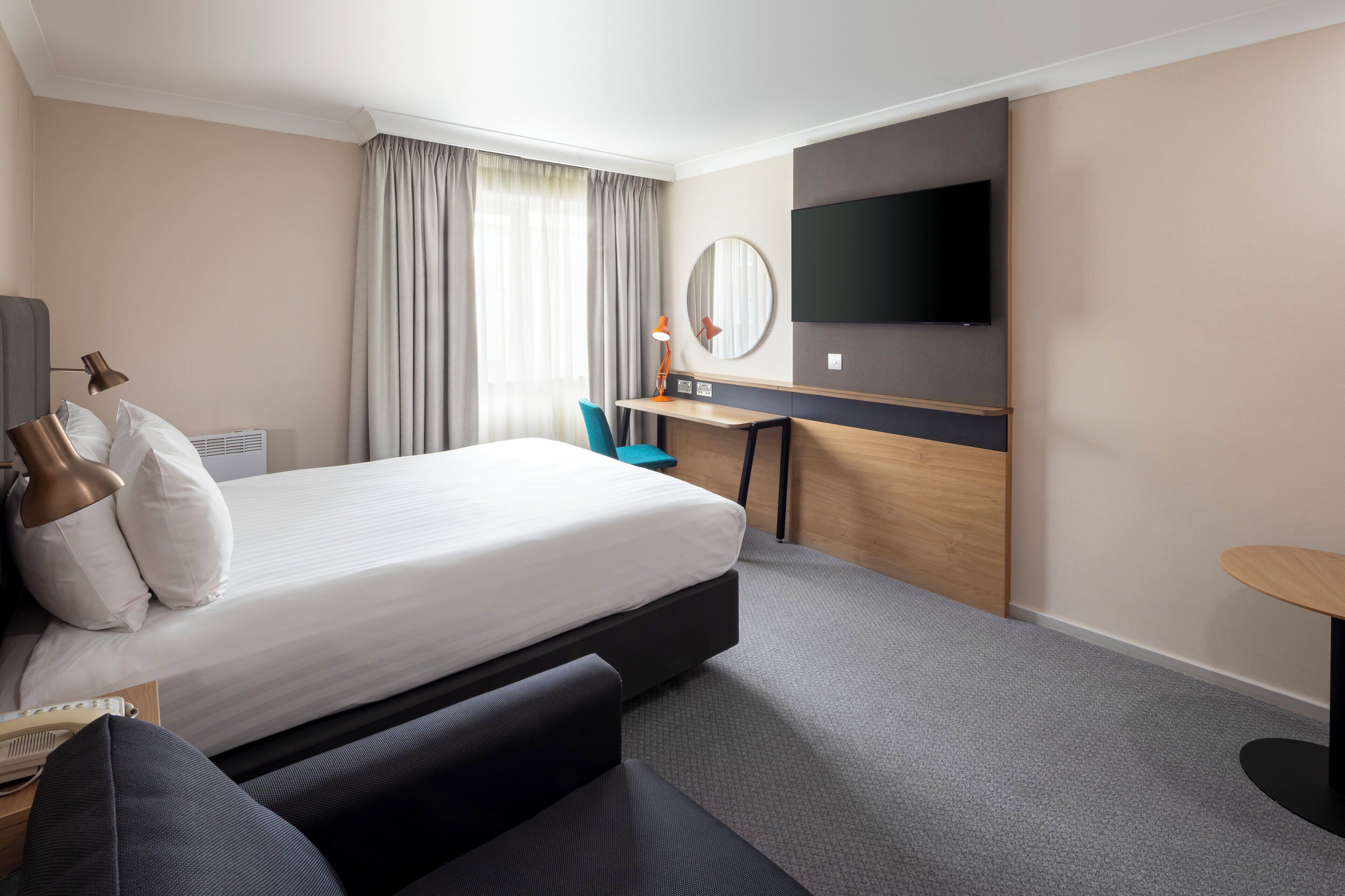 Una habitació d'hotel amb un llit, escriptori, cadira, televisió i una taula petita.