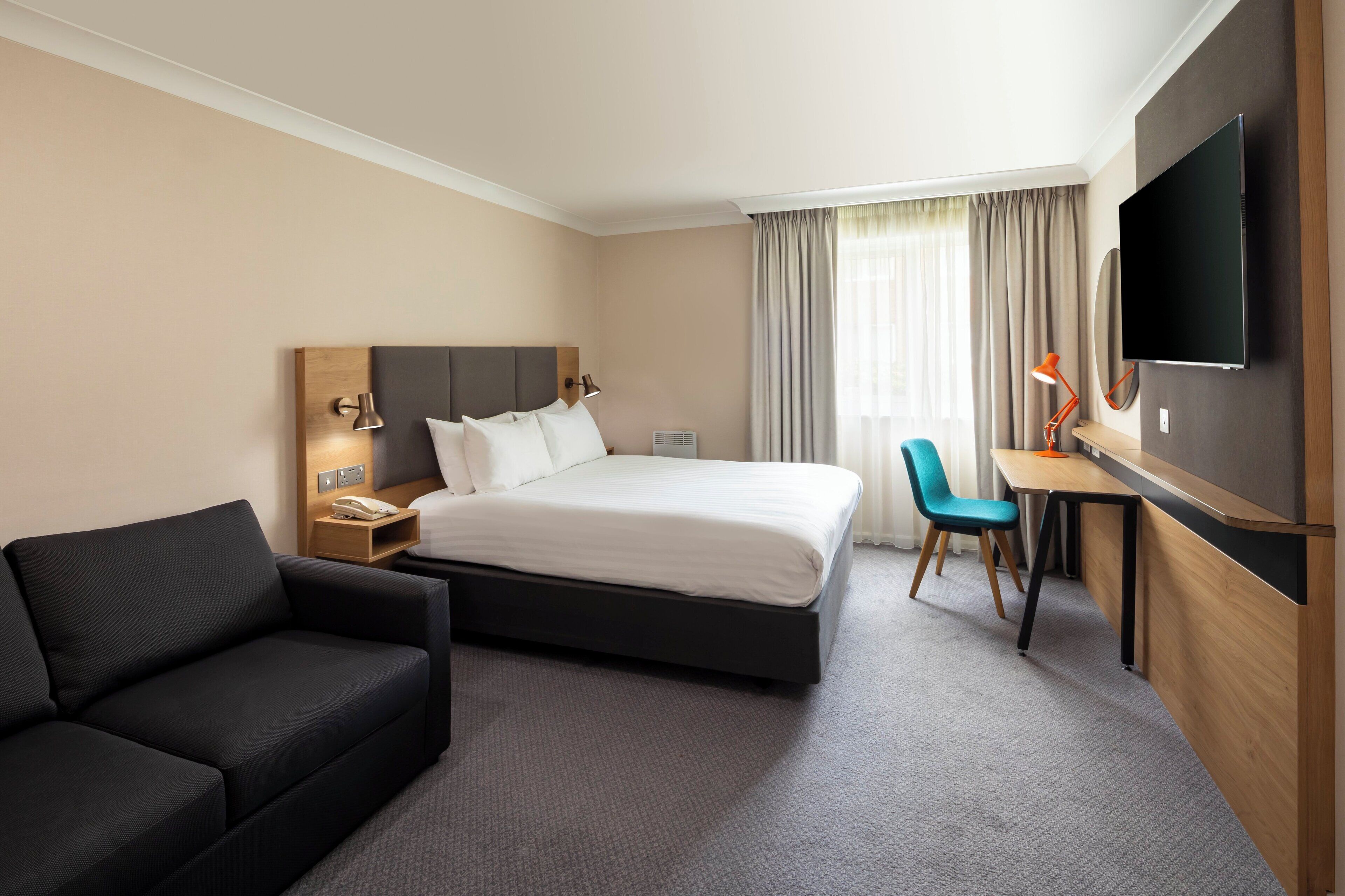 Habitación de hotel con cama, escritorio, silla, sofá, televisor y ventana con cortinas.