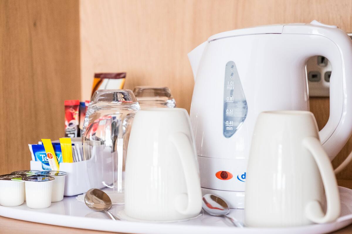 A white kettle, two white mugs, a glass pitcher, a spoon, and a container with tea packets on a white tray.