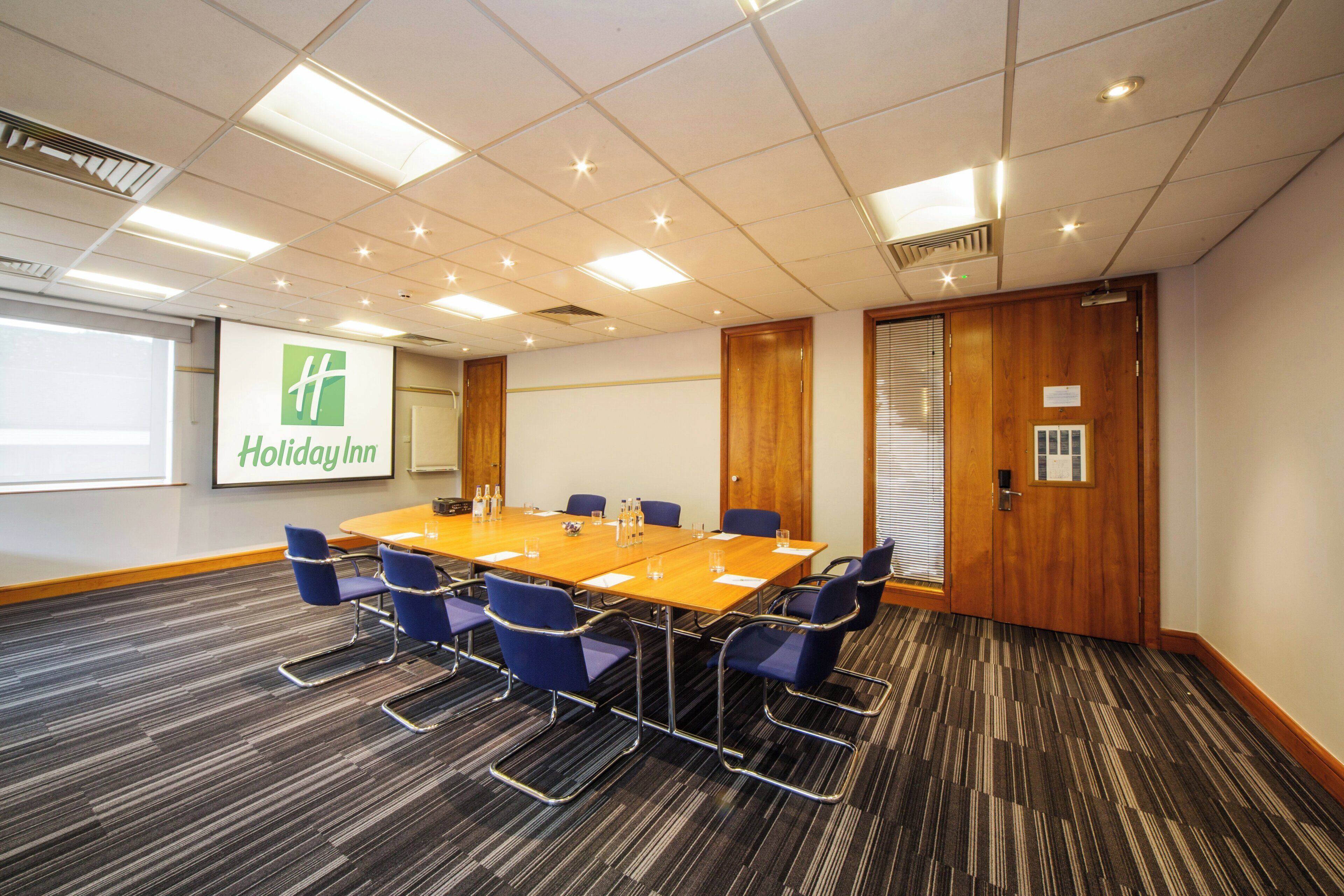 A conference room with a large table, blue chairs, a projector screen displaying 'Holiday Inn', and a wooden door.