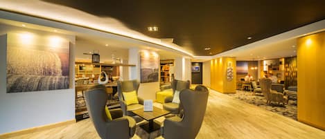 Un moderno vestíbulo de hotel con piso de madera, una zona de estar con sillones y una mesa de centro, una recepción y un cuadro grande en la pared.