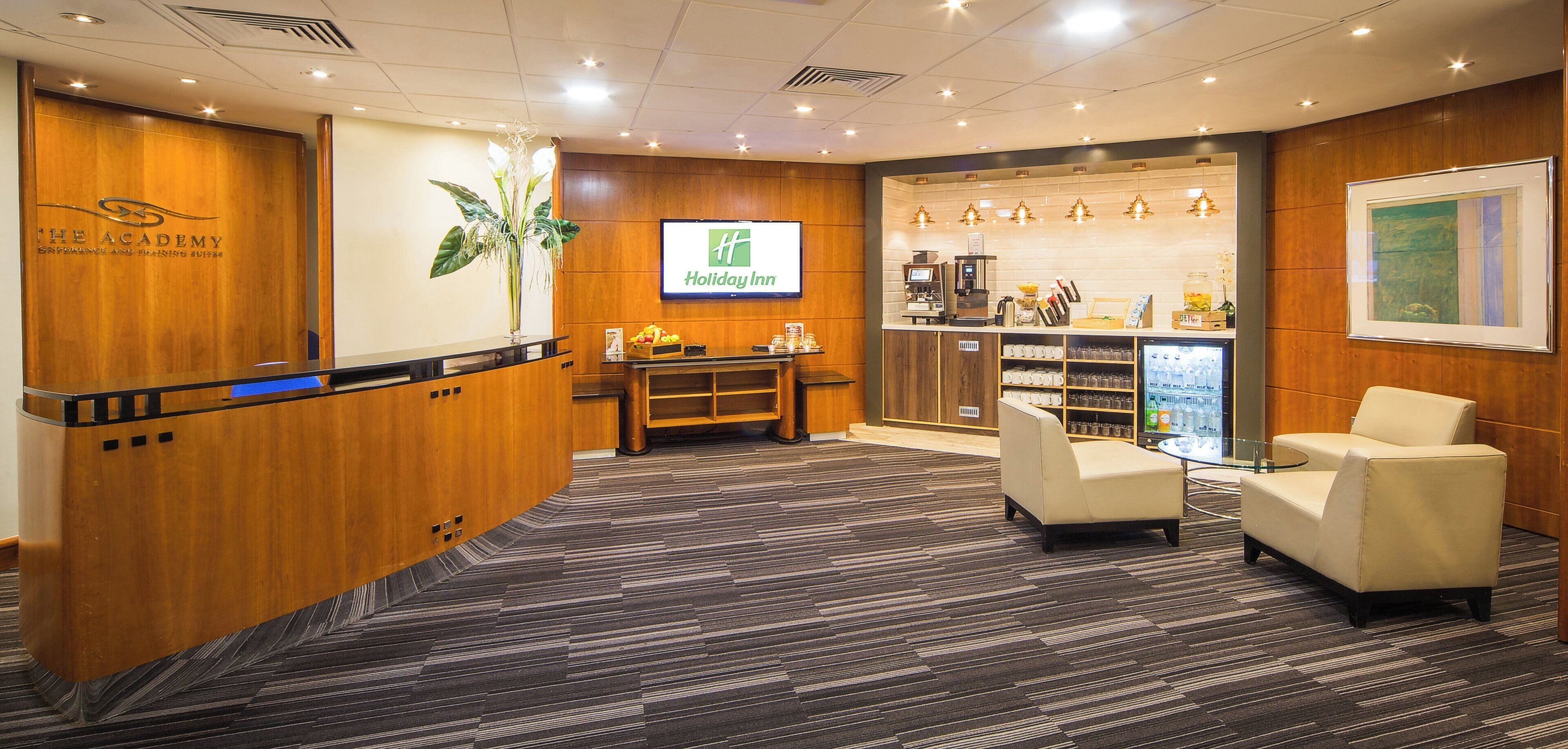 A hotel reception area with a wooden reception desk, a Holiday Inn sign, a coffee machine, a refrigerator, and seating area.