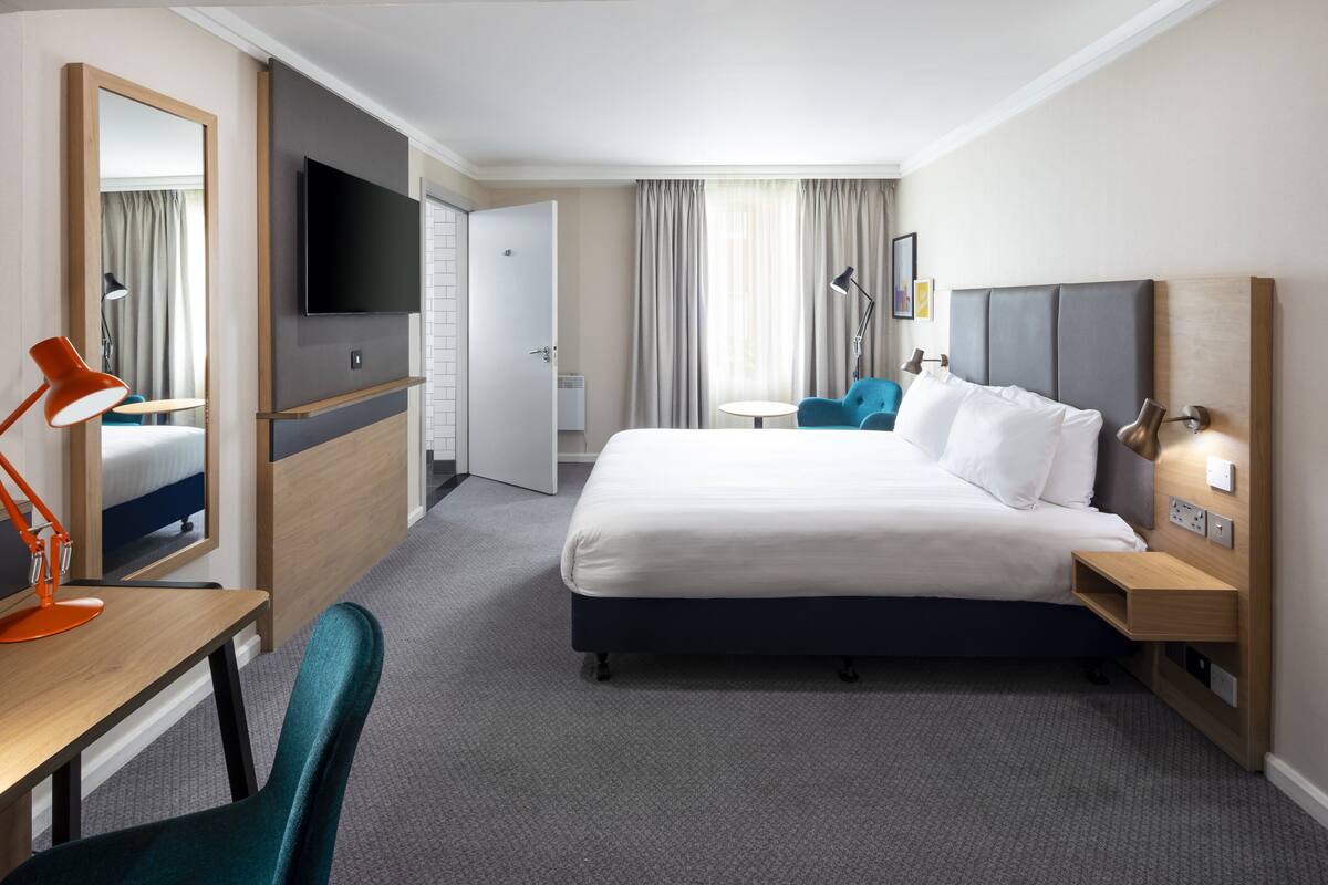 A modern hotel room with a large bed, a desk with a chair, a TV, and a small table with a chair.
