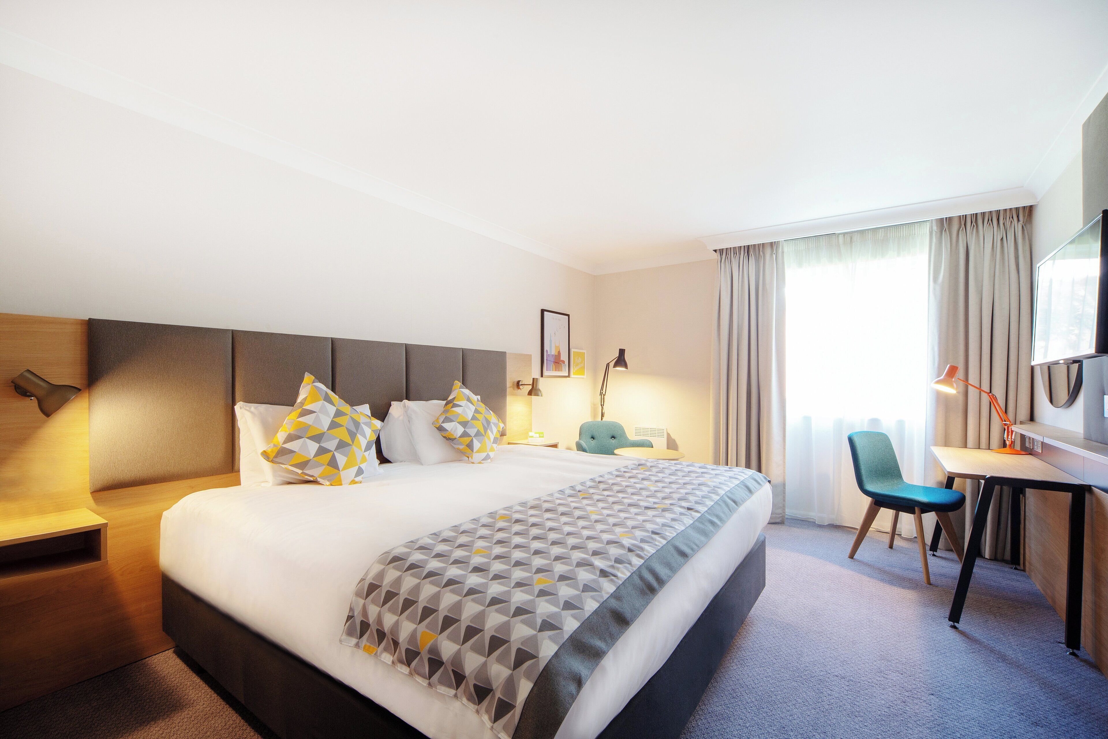 Une chambre d’hôtel avec un lit, un bureau, une chaise, une télévision et une fenêtre avec des rideaux.