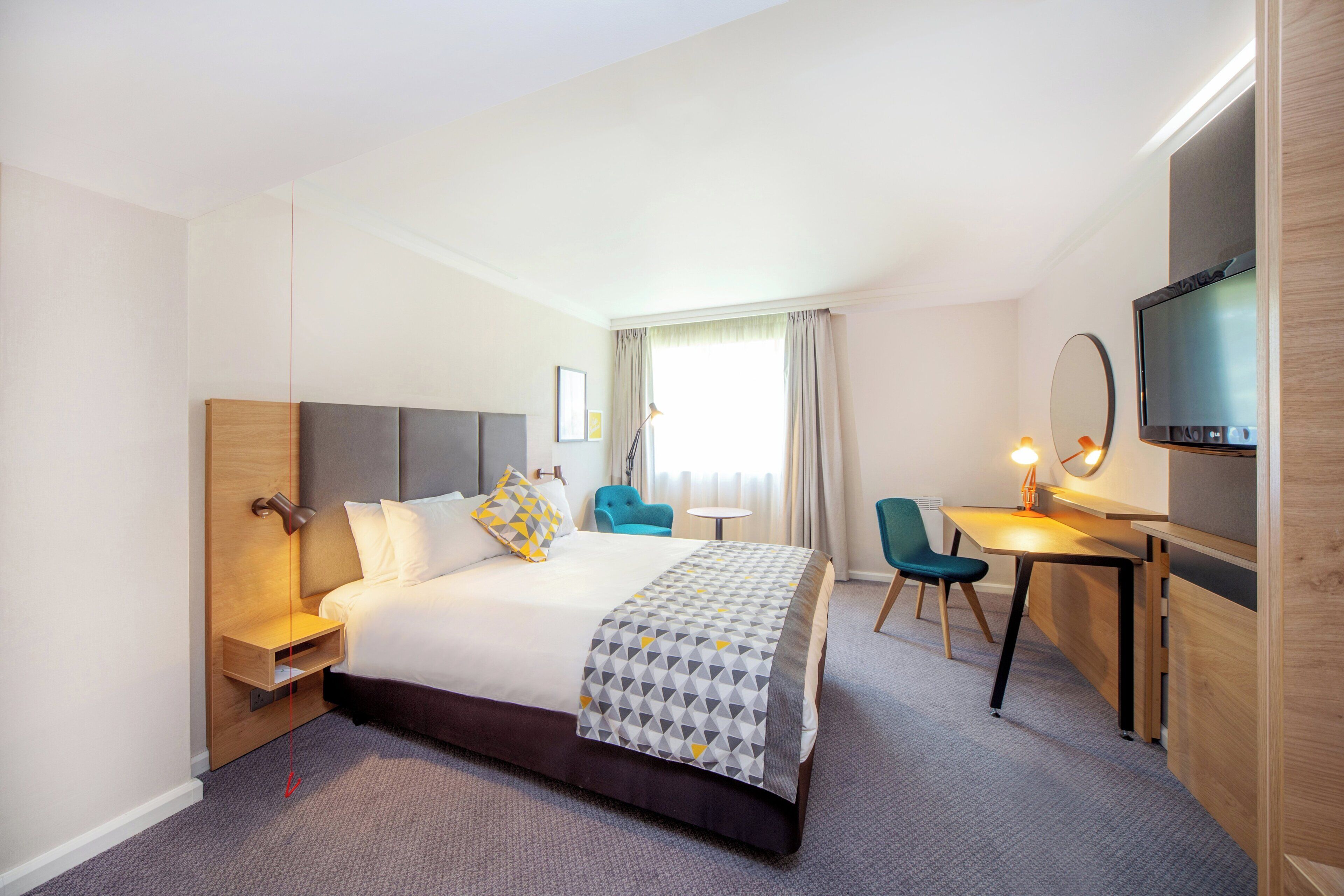 Ein Hotelzimmer mit Bett, Schreibtisch, Stuhl, Fernseher und einem Fenster mit Vorhängen.