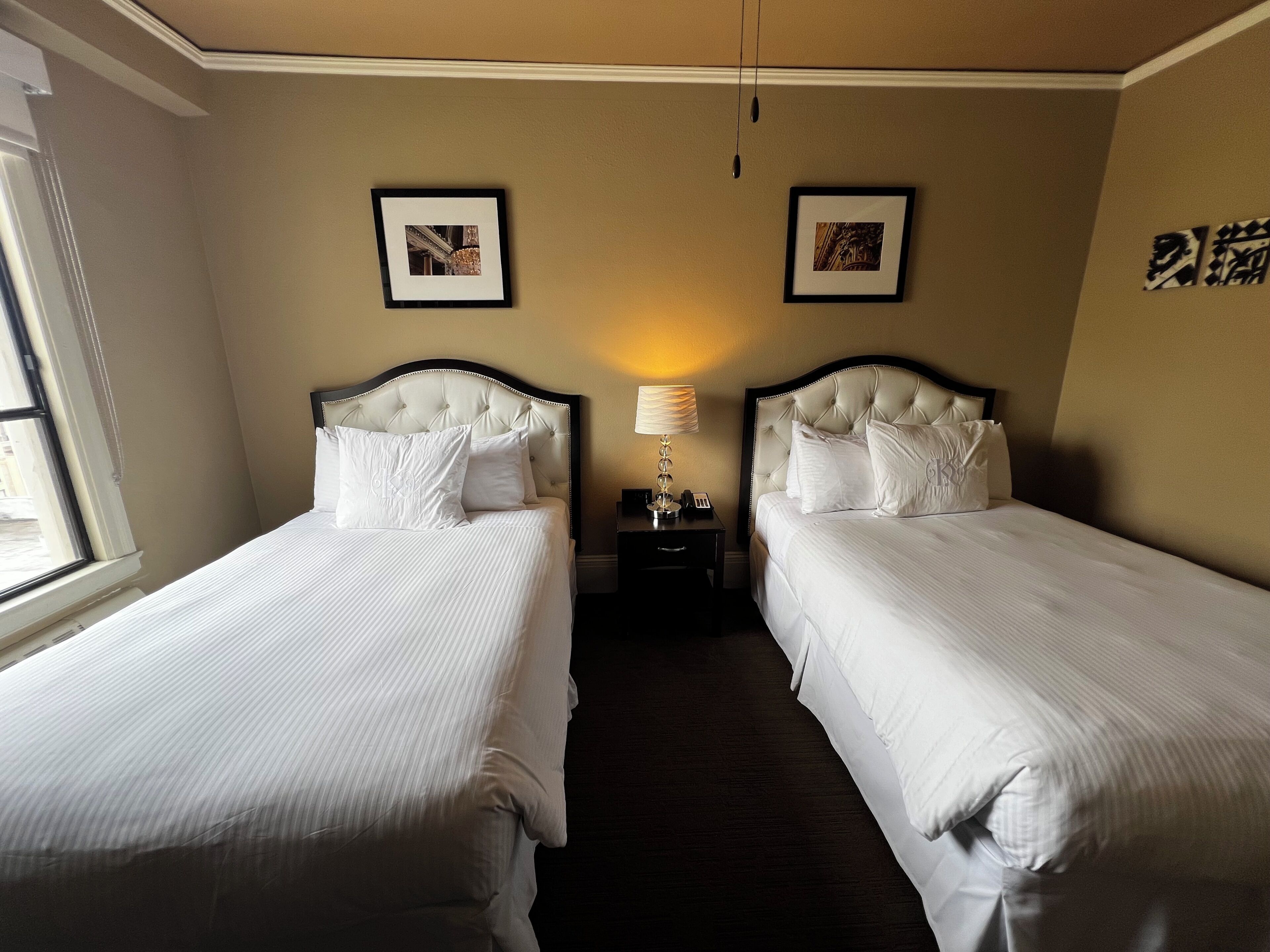 A hotel room with two beds, a nightstand, a lamp, and two framed pictures on the wall.