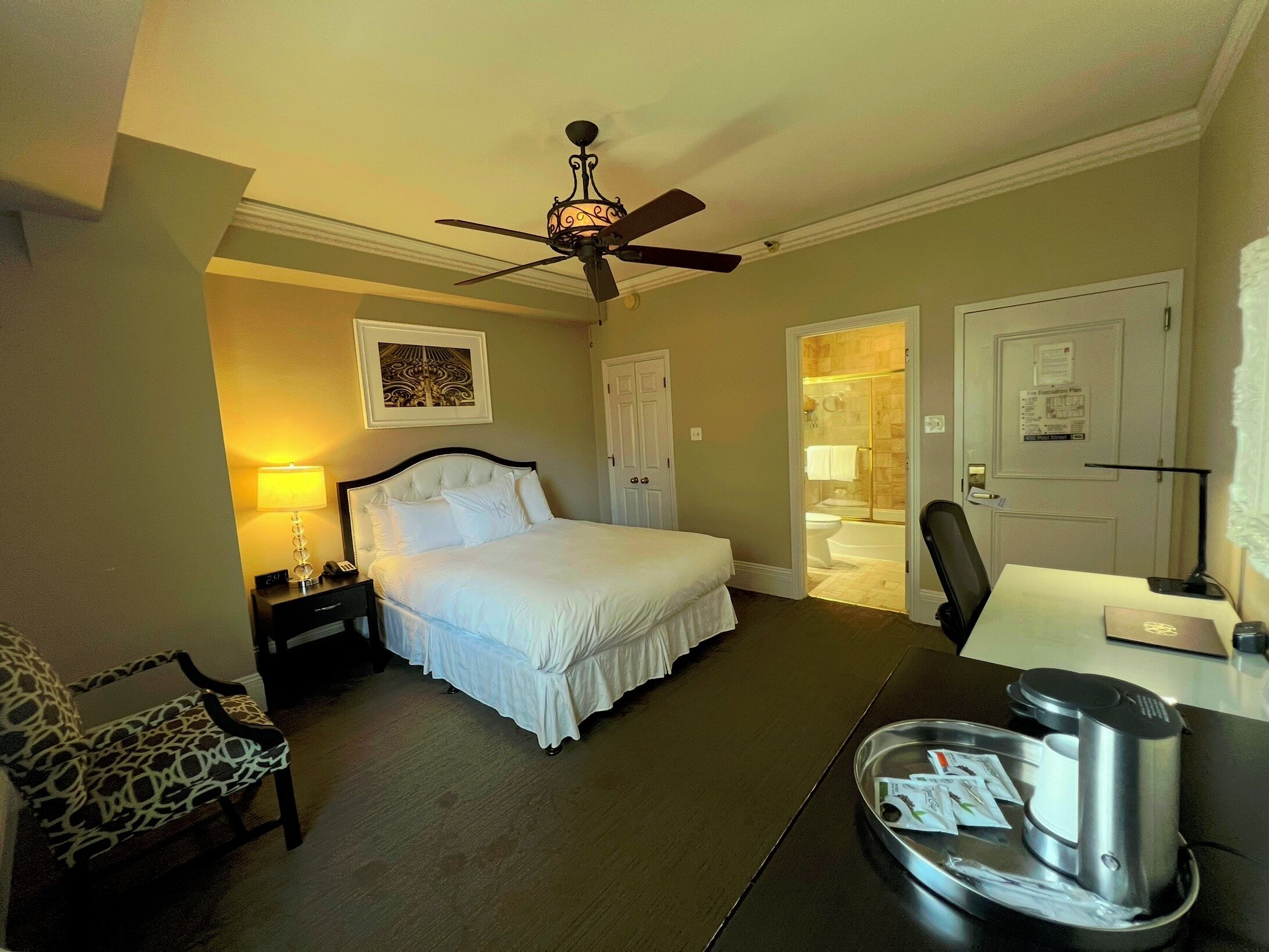A hotel room with a bed, a desk, a chair, a ceiling fan, and a bathroom door.