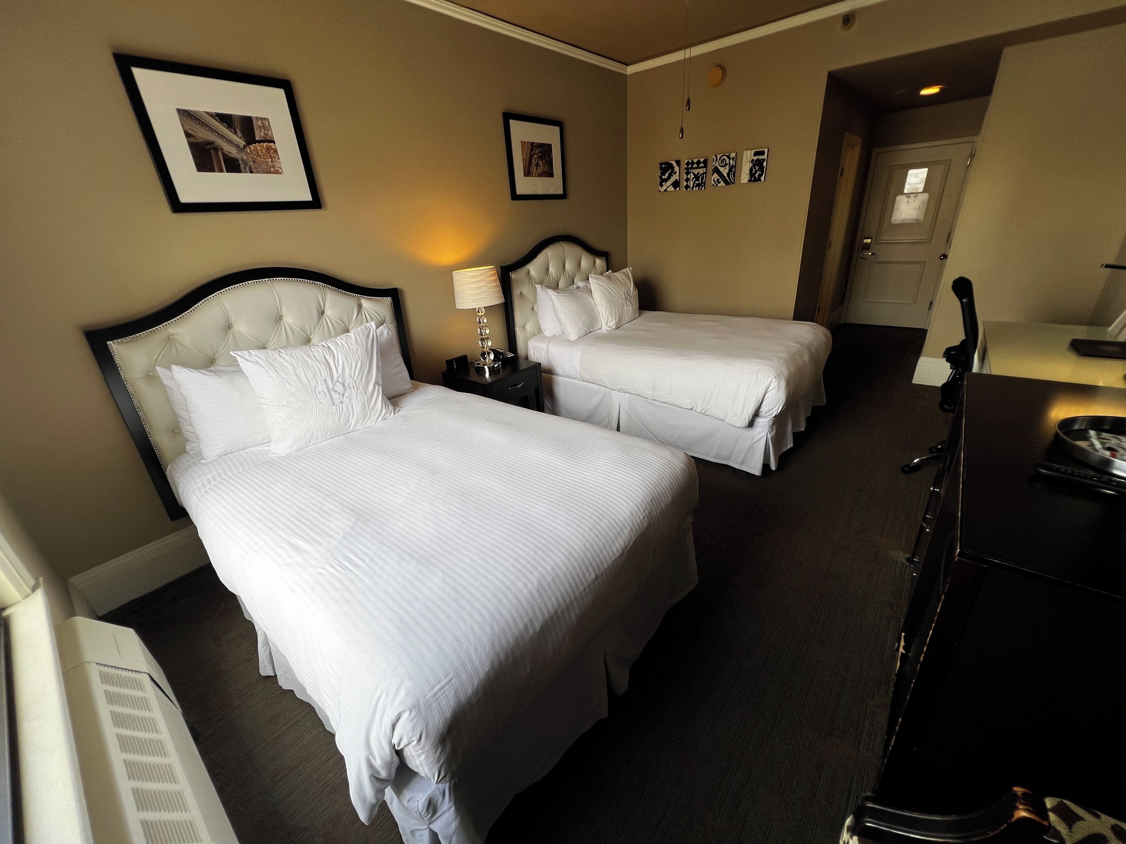 A hotel room with two beds, a dresser, a lamp, and framed pictures on the wall.