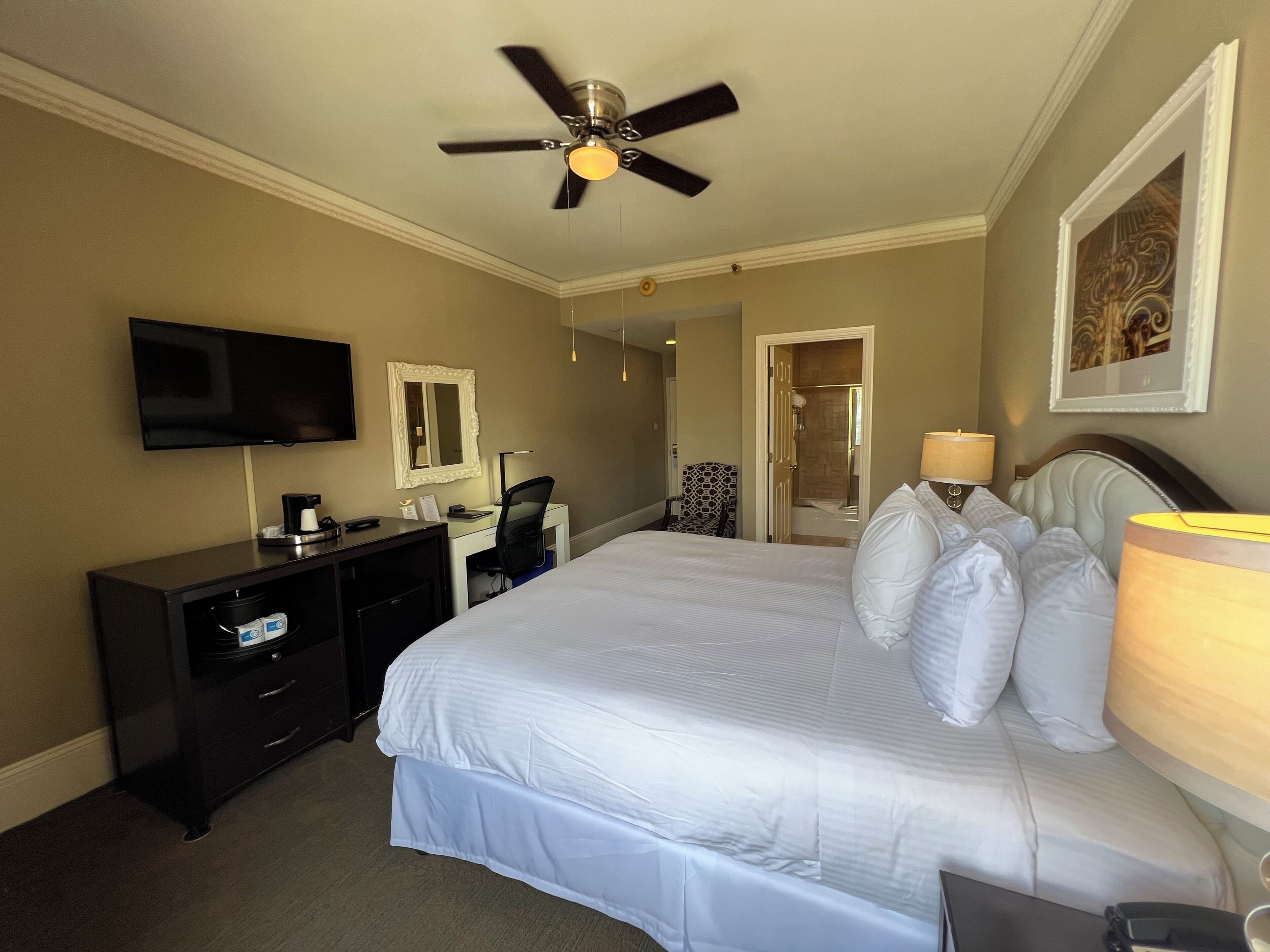 A hotel room with a bed, a desk, a chair, a television, a ceiling fan, and a door leading to a bathroom.