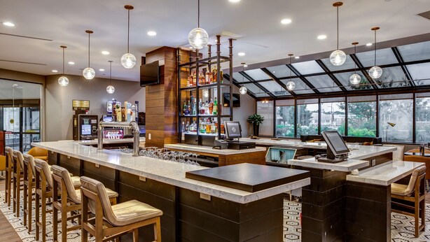 Een moderne bar met een lange toonbank, diverse barkrukken en een assortiment flessen op planken.
