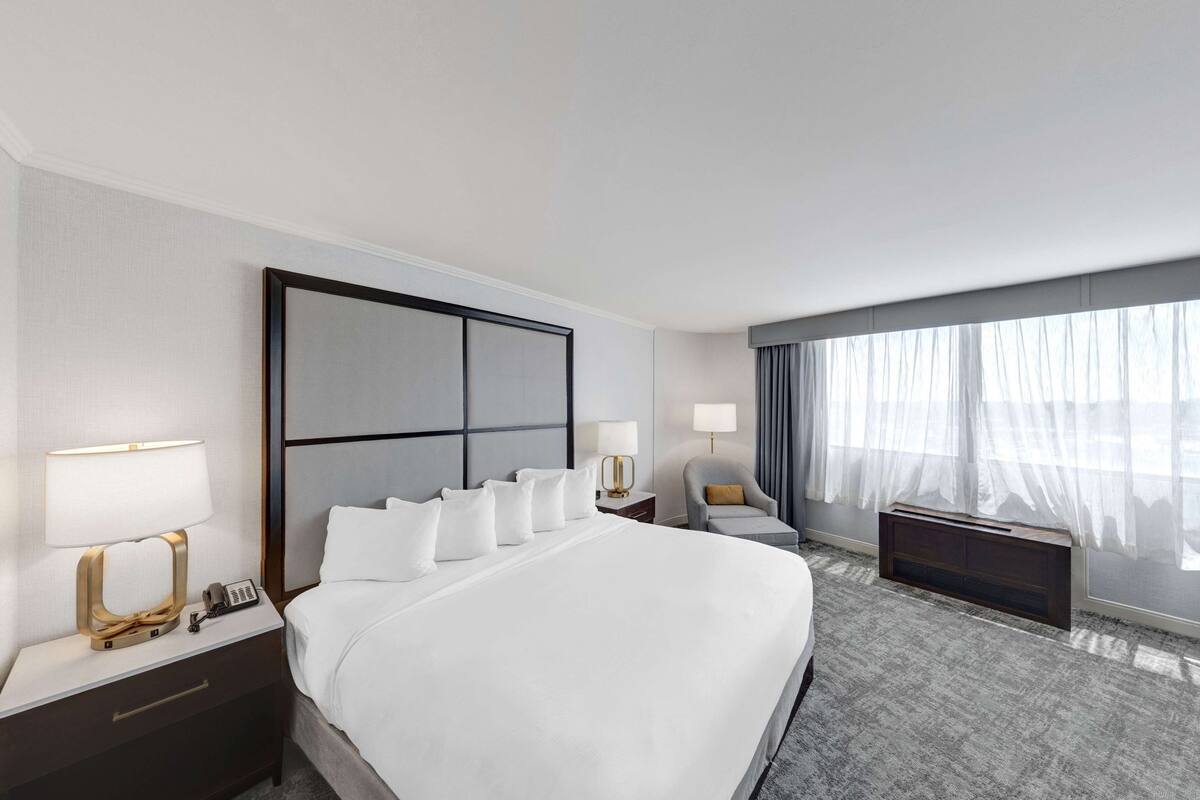 A hotel room with a large bed, bedside tables with lamps, a chair, a desk, and a large window with curtains.