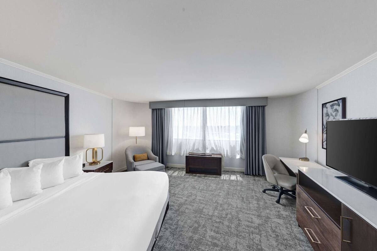 A hotel room with a large bed, a desk with a flat-screen TV, a chair, a small table, and a window with curtains.