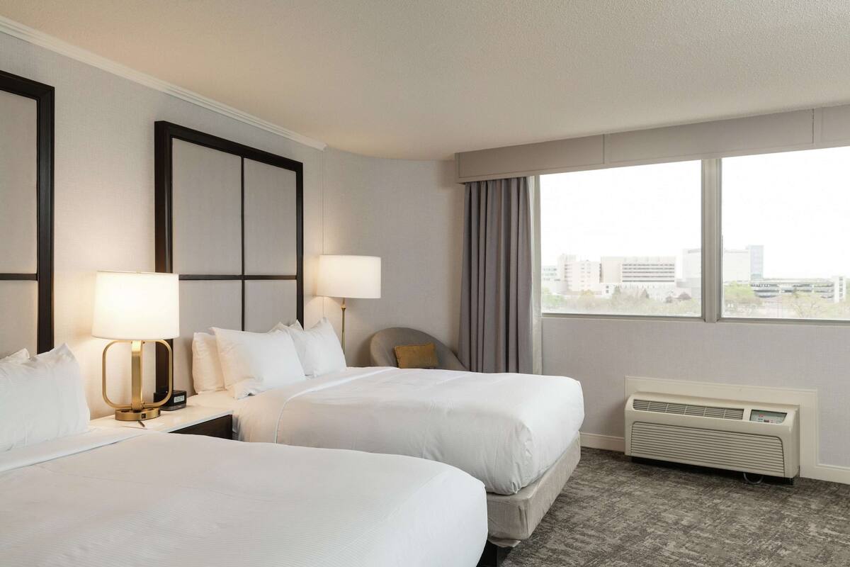 A hotel room with two beds, a bedside table with a lamp, a window with curtains, and a wall-mounted air conditioner.