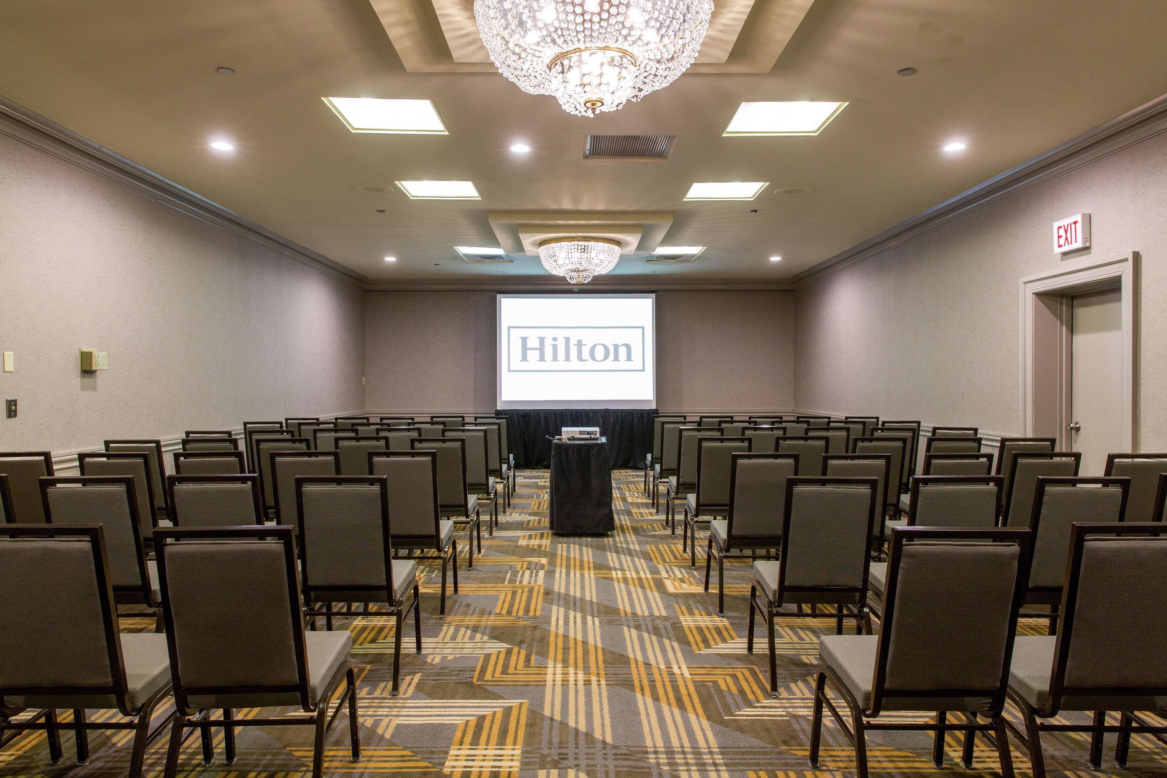 Un salón de conferencias con filas de sillas, un atril y una pantalla de proyector que muestra "Hilton".