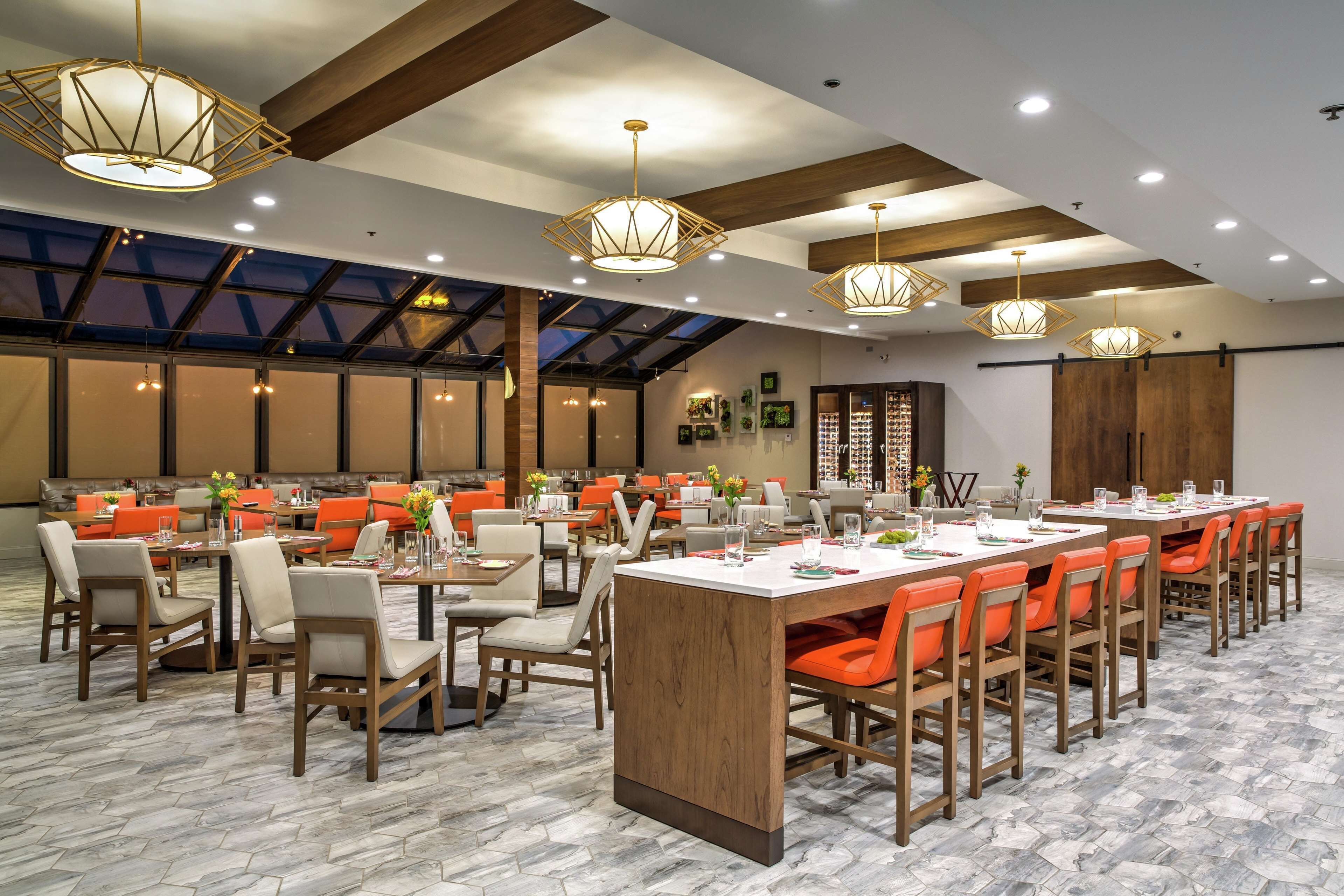 A spacious dining area with multiple tables and chairs, featuring a modern design with wooden accents and orange chairs.