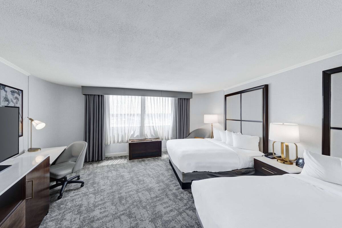 A hotel room with a large flat-screen TV, a desk with a chair, two beds, a nightstand with a lamp, and a large window with curtains.