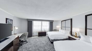 A hotel room with a large flat-screen TV, a desk with a chair, two beds, a nightstand with a lamp, and a large window with curtains.