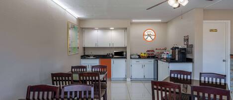 A dining area with a table and chairs, a kitchen in the background, and a ceiling fan.