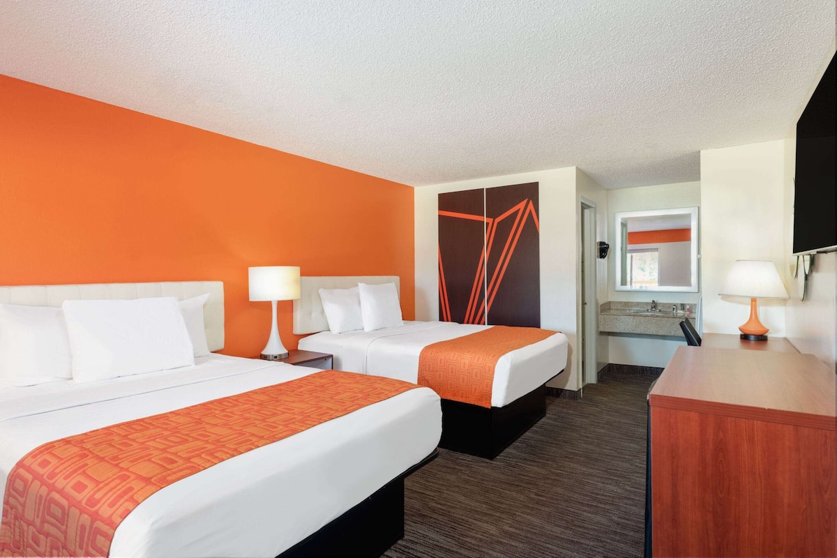 A hotel room with two beds, a desk, a mirror, and a bathroom visible through an open door.