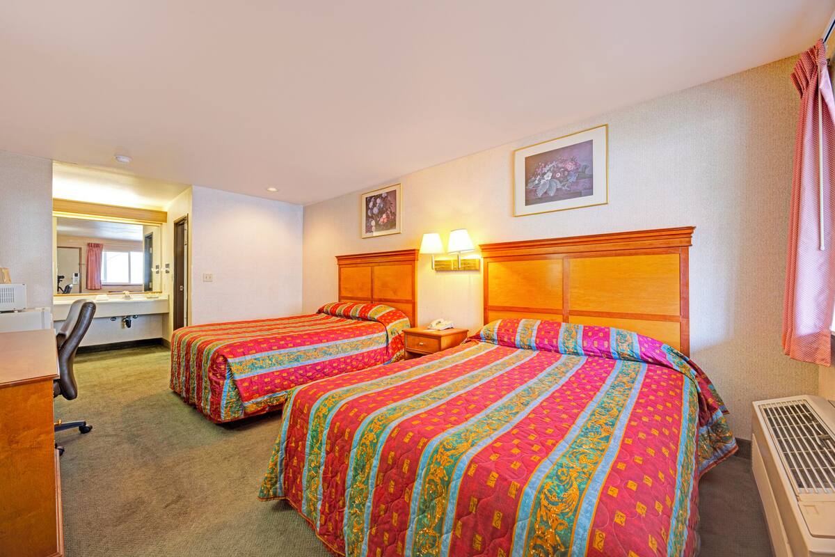 A hotel room with two beds, a desk, a chair, a nightstand, a lamp, and a window with a curtain.