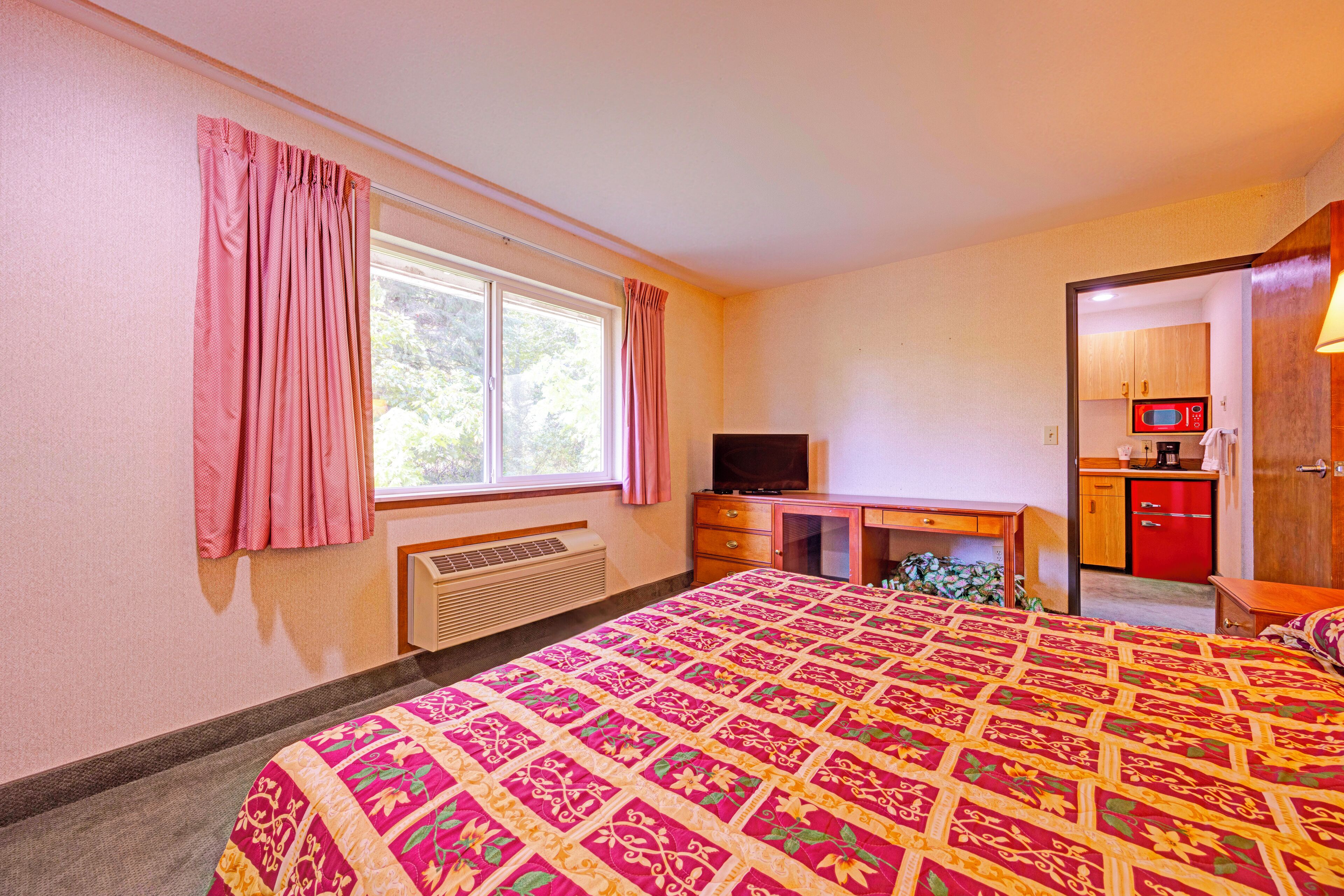 A bedroom with a large bed, a desk, a television, and a kitchenette.