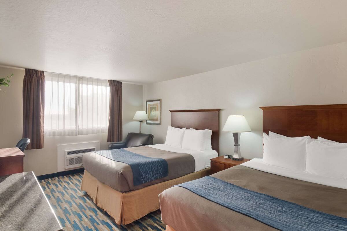 A hotel room with two beds, a desk, a chair, a lamp, and a window with curtains.