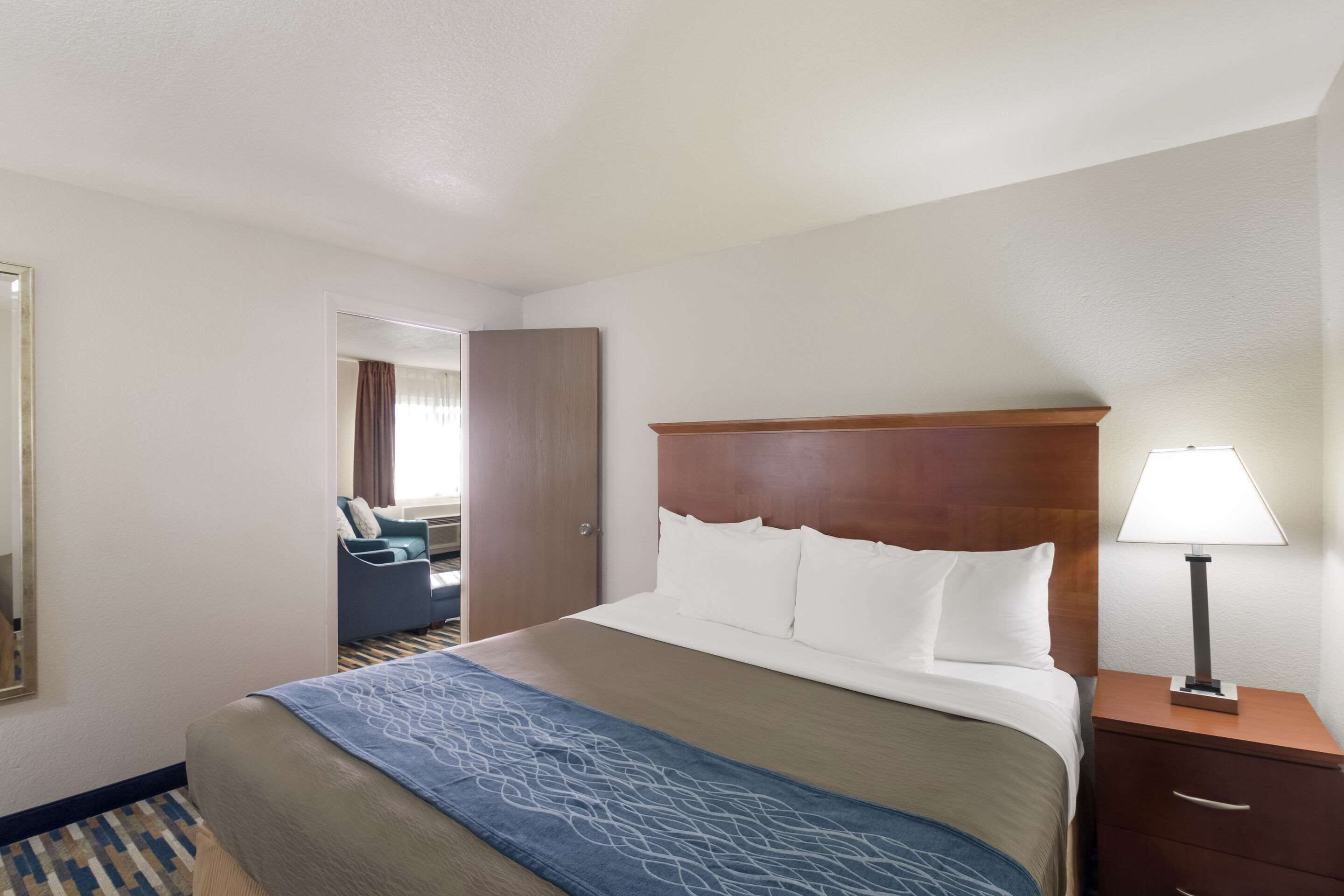 A hotel room with a bed, a nightstand, a lamp, a sofa, and a door leading to another room.
