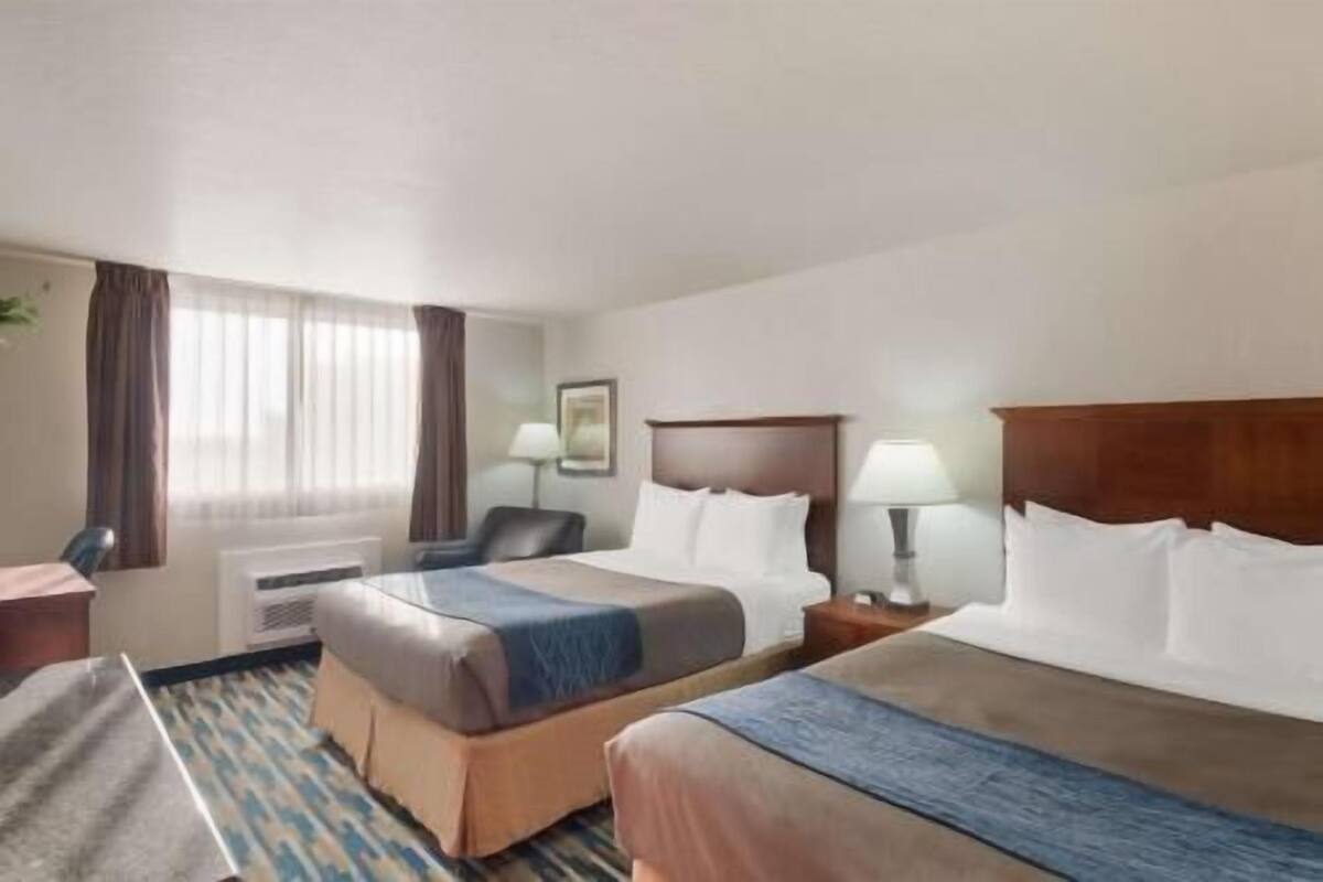 A hotel room with two beds, a desk, a chair, a lamp, and a window with curtains.