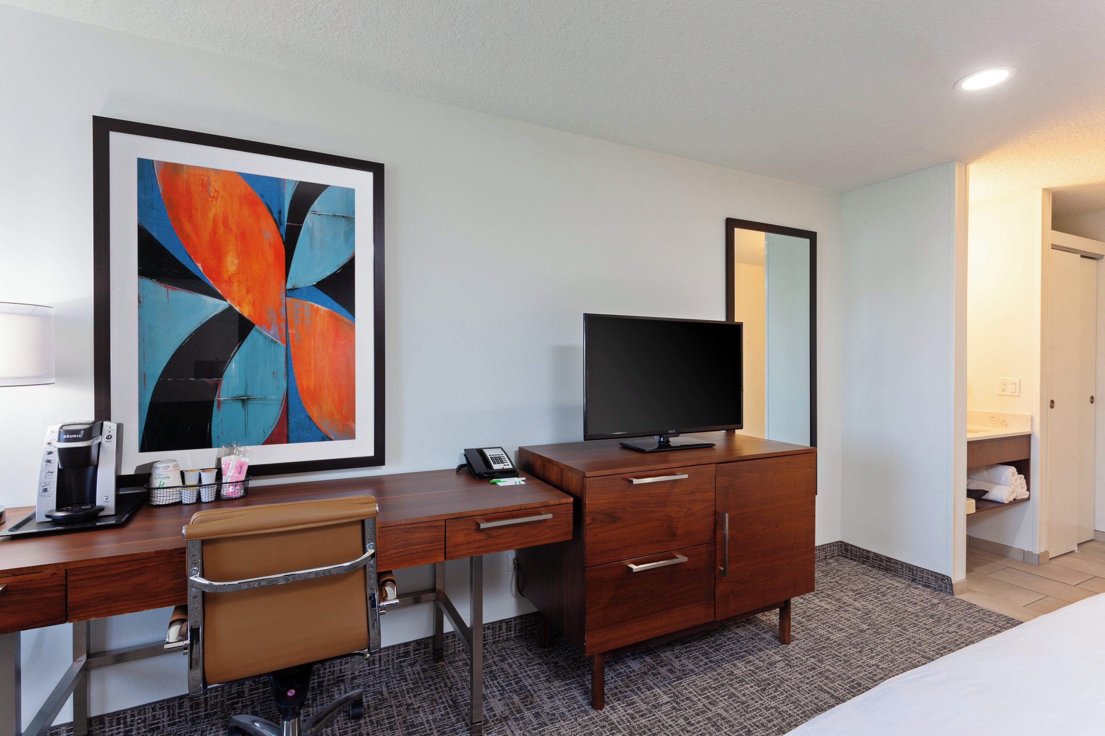 Habitación de hotel con escritorio, silla, cafetera, teléfono, televisor y un cuadro abstracto grande.