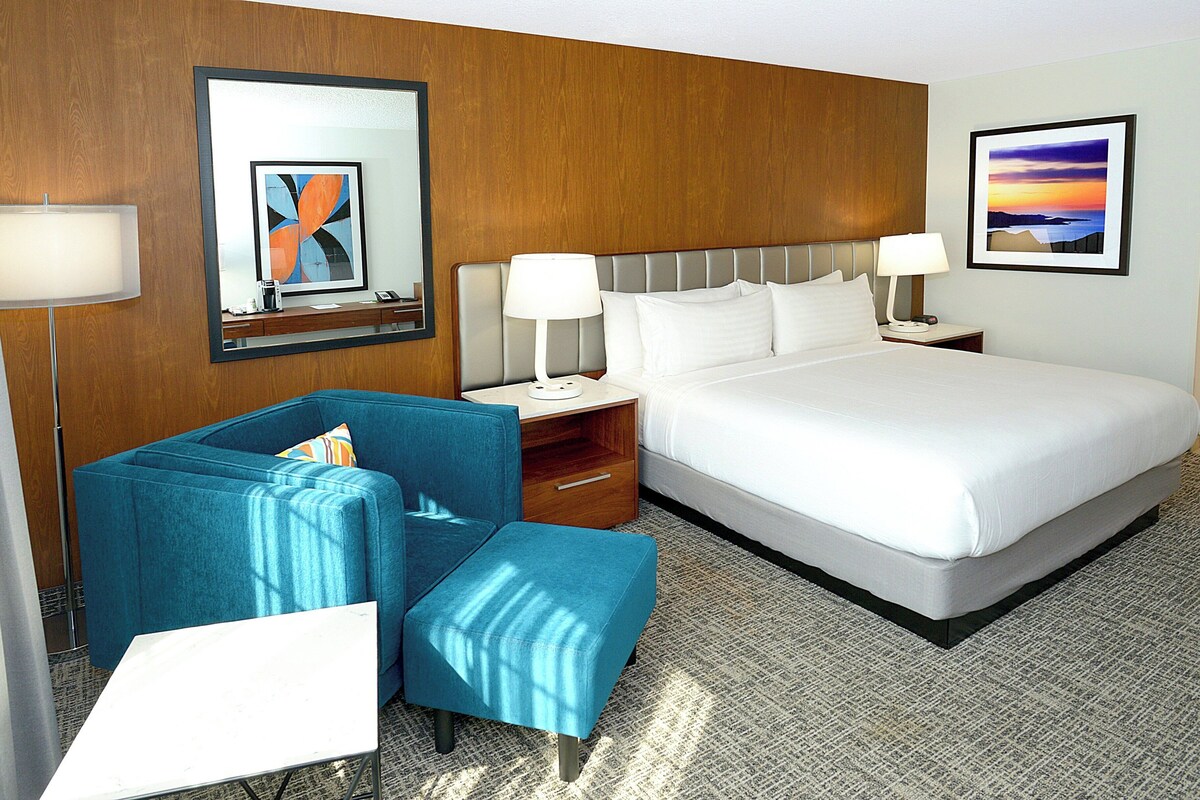 Een hotelkamer met een groot bed, een blauwe fauteuil, een klein tafeltje, een nachtkastje en twee wandlampen.