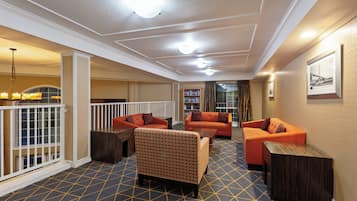 Un vestíbulo de hotel con alfombra estampada, sofás naranja, un sillón con diseño, una mesa de centro de madera y una estantería.