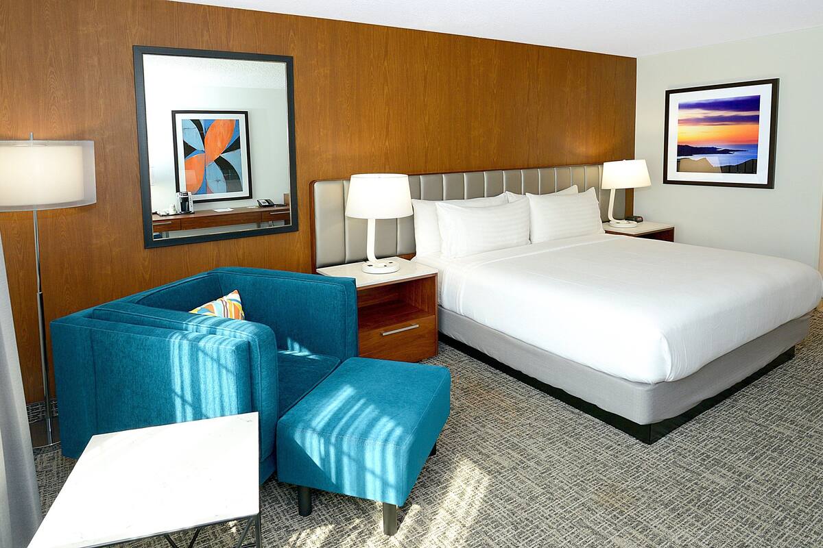 Een hotelkamer met een groot bed, een blauwe fauteuil, een kleine tafel en een nachtkastje met lampen.
