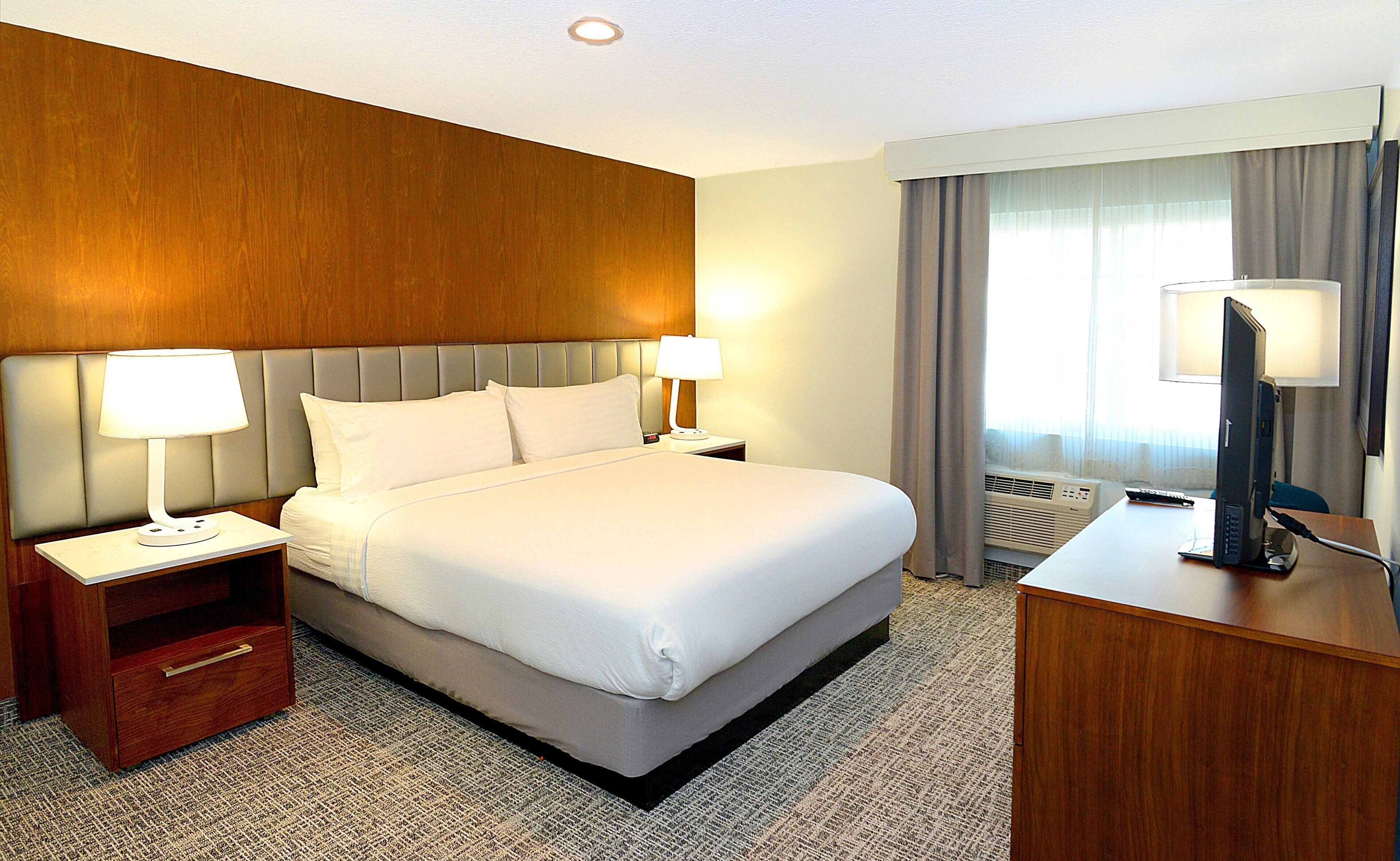 Quarto de hotel com uma cama grande, duas mesas de cabeceira com luminárias, uma escrivaninha com computador e uma janela com cortinas.