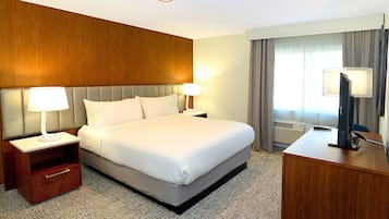 Quarto de hotel com uma cama grande, duas mesas de cabeceira com luminárias, uma escrivaninha com computador e uma janela com cortinas.