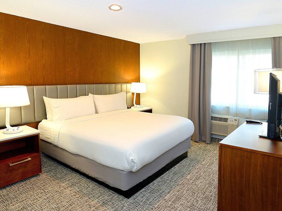 Quarto de hotel com uma cama grande, duas mesas de cabeceira com luminárias, uma escrivaninha com computador e uma janela com cortinas.