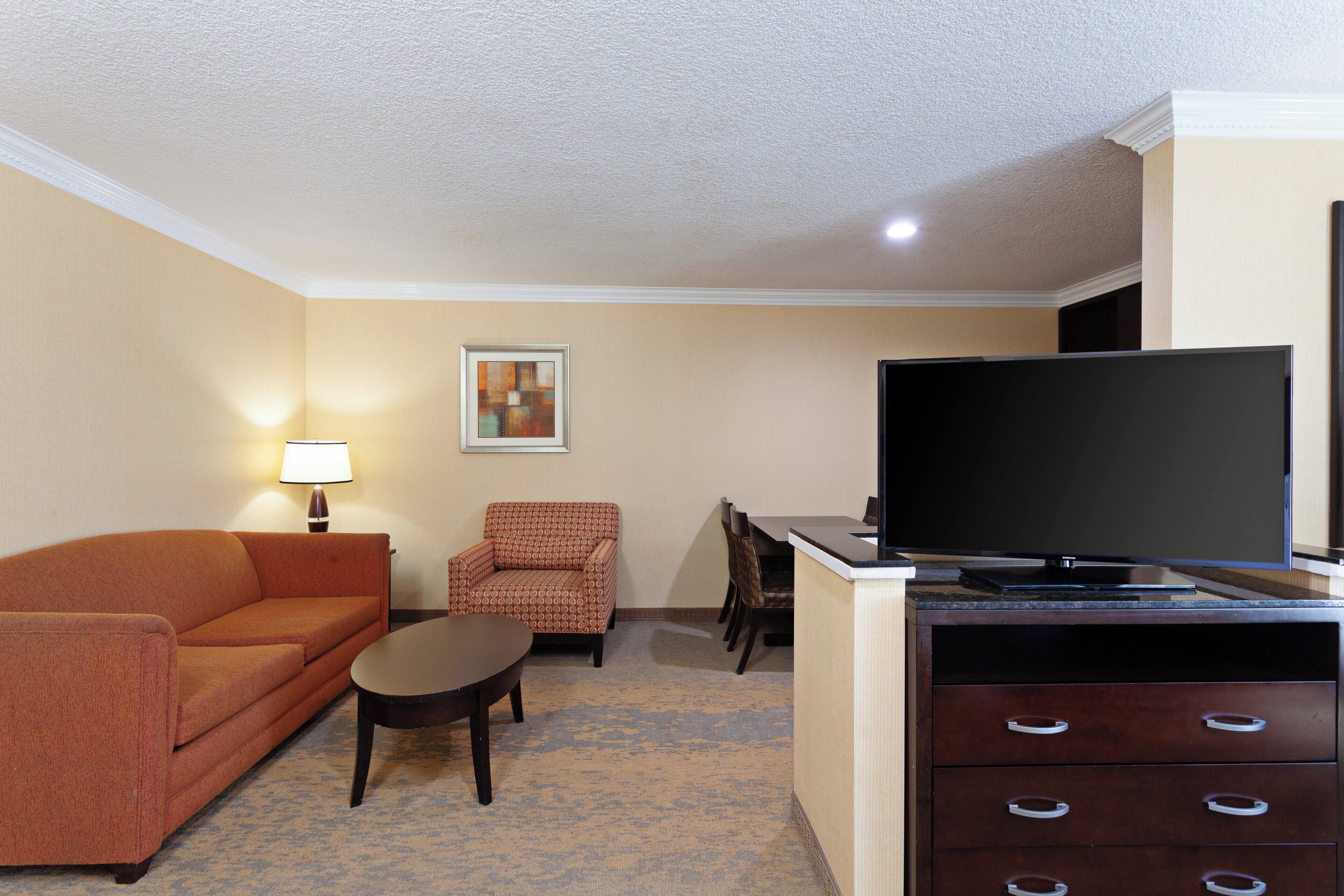 A hotel room with a sofa, armchair, dining table, chairs, a flat-screen TV, and a cabinet.