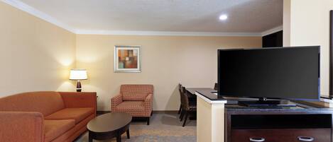 A hotel room with a sofa, armchair, dining table, chairs, a flat-screen TV, and a cabinet.